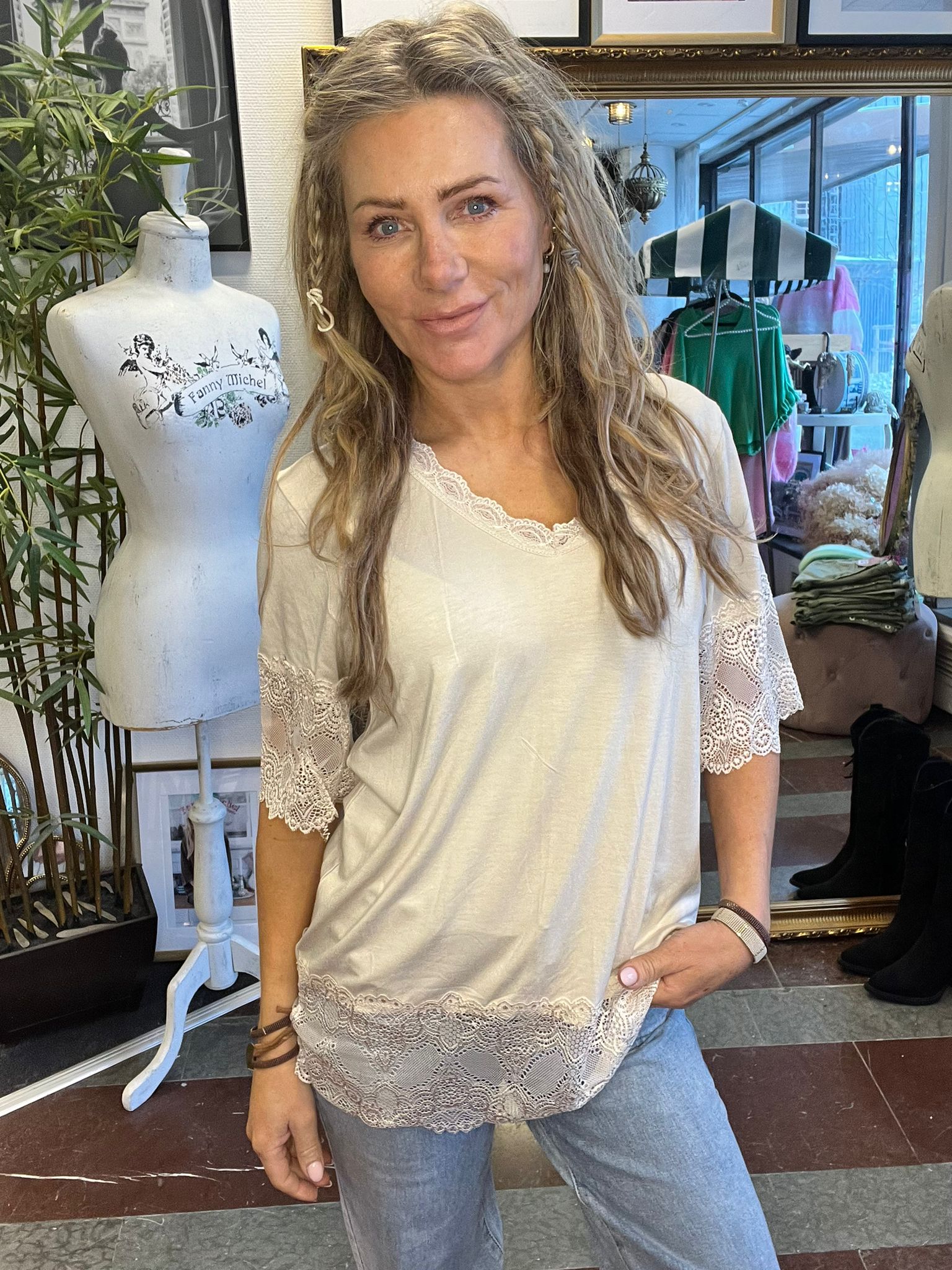 Kvinna står och ler mot kameran i en klädbutik, med handen i jeansfickan och en vit topp med spetsdetaljer. Bredvid syns en provdocka med texten “Fanny Michel” samt kläder och inredning i bakgrunden.
