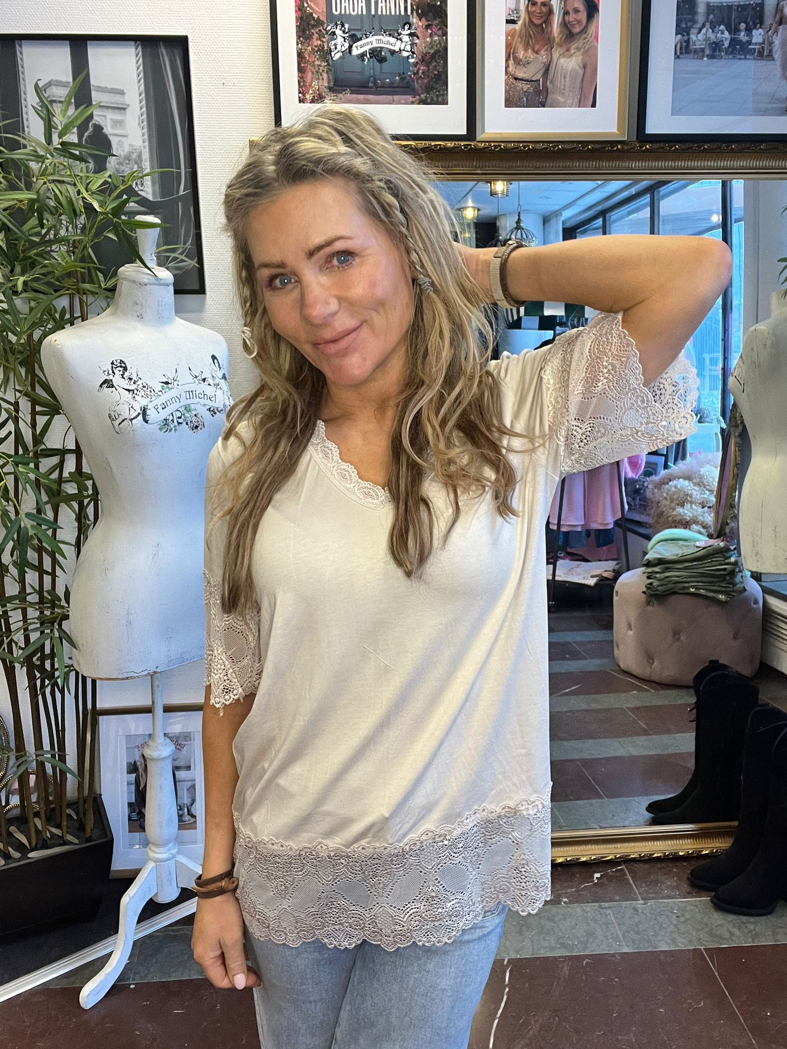 Blond kvinna poserar och ler med ena armen bakom huvudet, klädd i vit tunika med spets. Hon står i en butik bredvid en provdocka med texten ”Fanny Michel”, med spegel och inramade foton i bakgrunden.