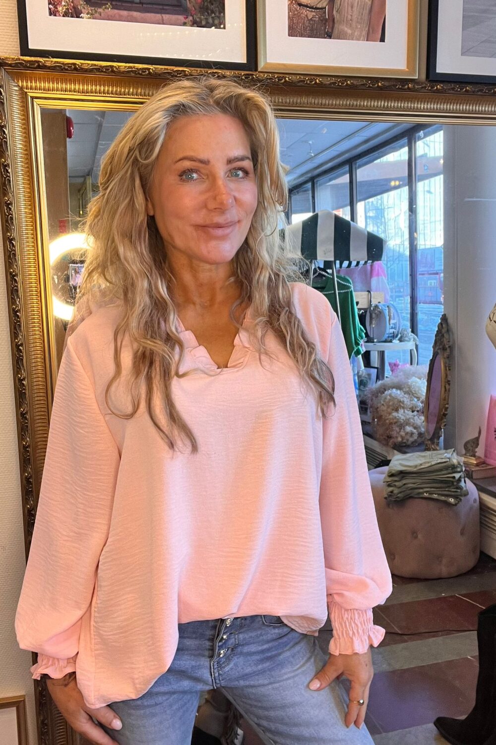 Leende kvinna med långt blont hår står och poserar i en ljusrosa blus och jeans framför en stor guldramsspegel. I bakgrunden syns butiksmiljö med kläder, provdocka, puff och stora fönster.