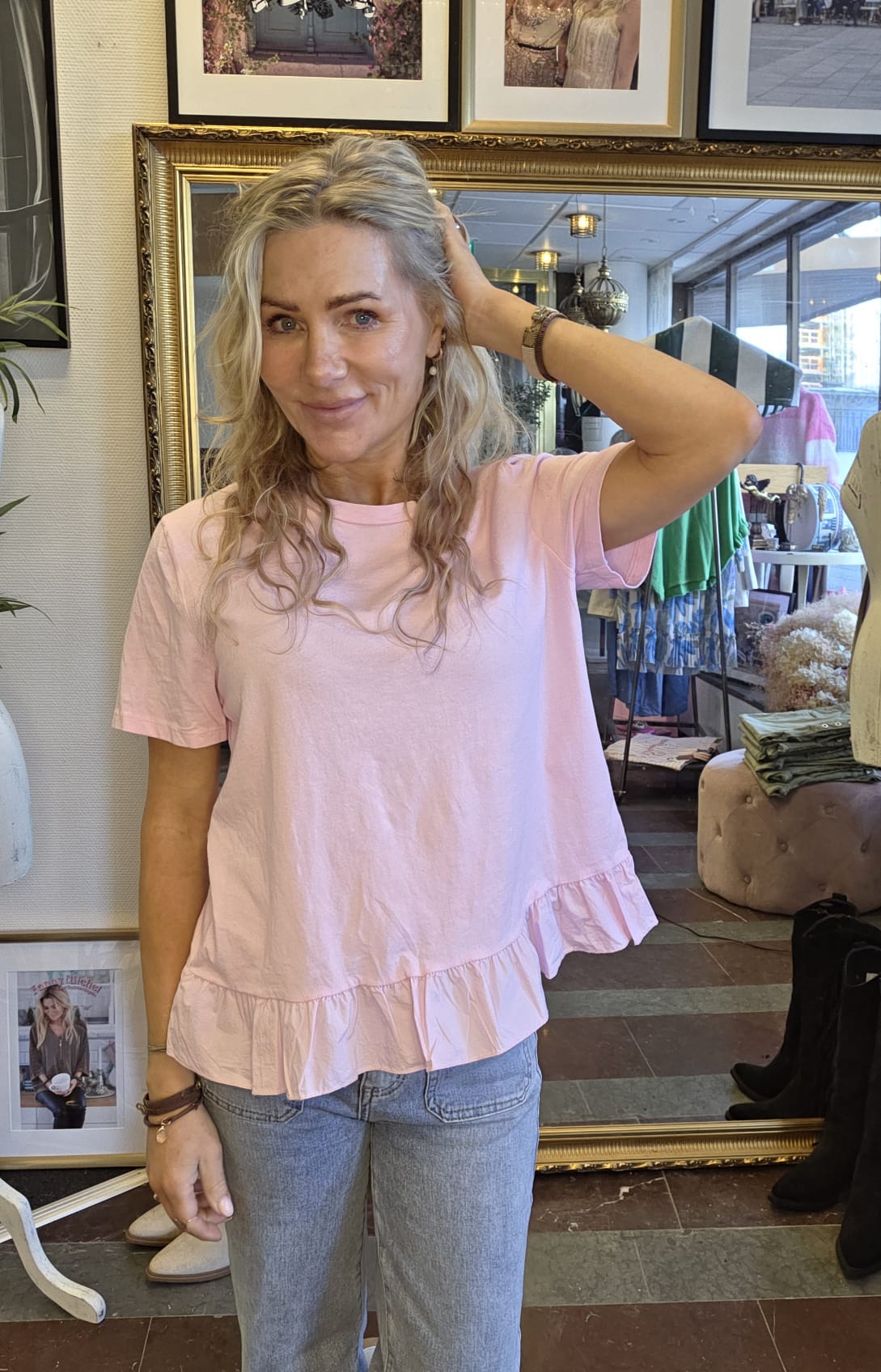 Kvinna med långt blont hår står och ler, med ena handen i håret. Hon bär ljusrosa topp med volangkant och jeans, i en butiksmiljö med stor guldramsspegel, klädställningar och inramade foton på väggen.