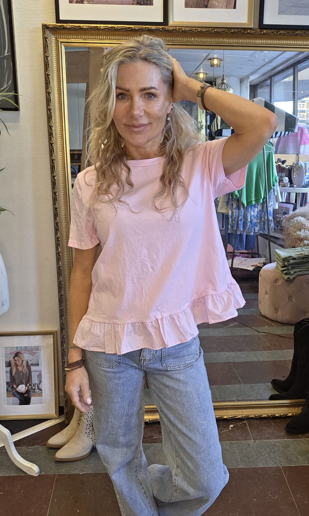 Kvinna med blont, vågigt hår poserar och håller handen i håret, klädd i ljusrosa volangblus och ljusa jeans. Hon står i en butik framför en stor guldramsspegel, med kläder och skor i bakgrunden.