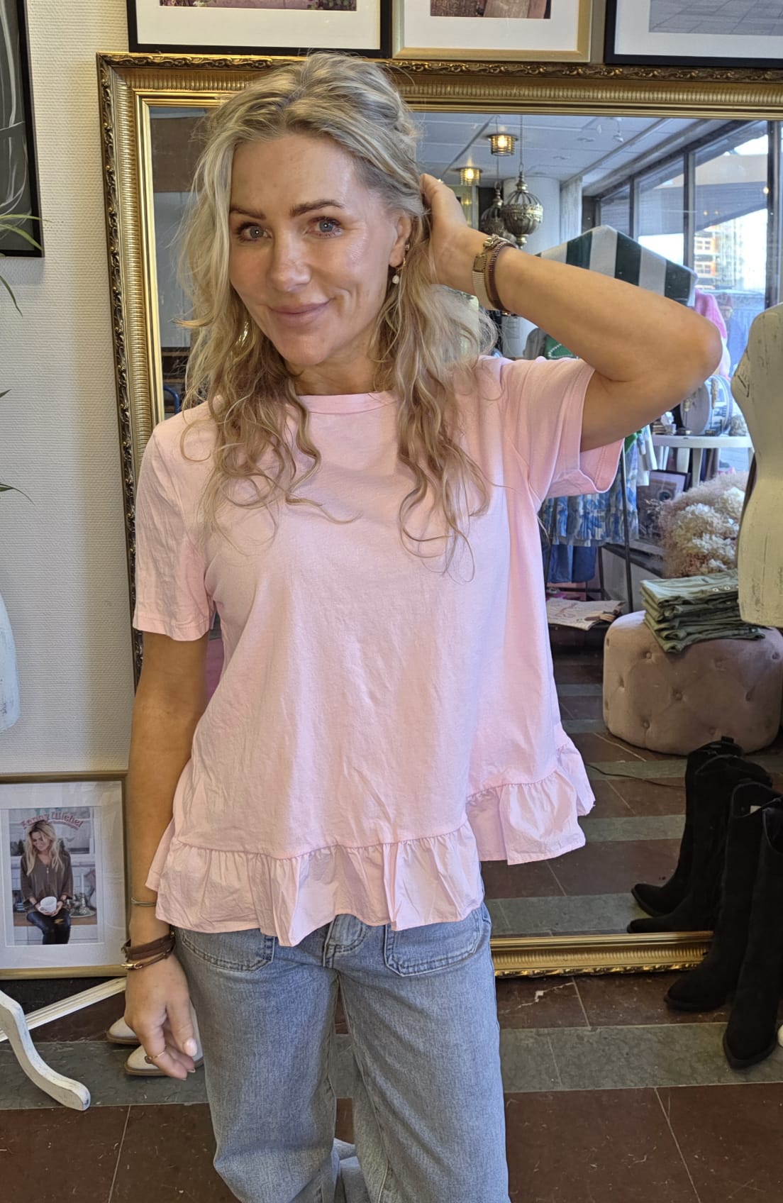 Blond kvinna i ljusrosa topp med volangkant och jeans står och ler, med ena handen bakom huvudet. Hon är inomhus i en butik med stor guldramsspegel, kläder och stövlar synliga i bakgrunden.