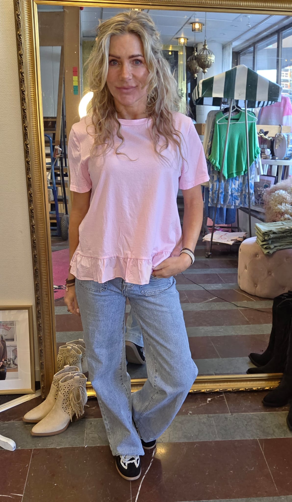 Kvinna med långt lockigt blont hår står framför en stor guldramsspegel och poserar med ena handen i jeansfickan. Hon bär ljusrosa topp med volangkant och ljusblå jeans i en klädbutik.