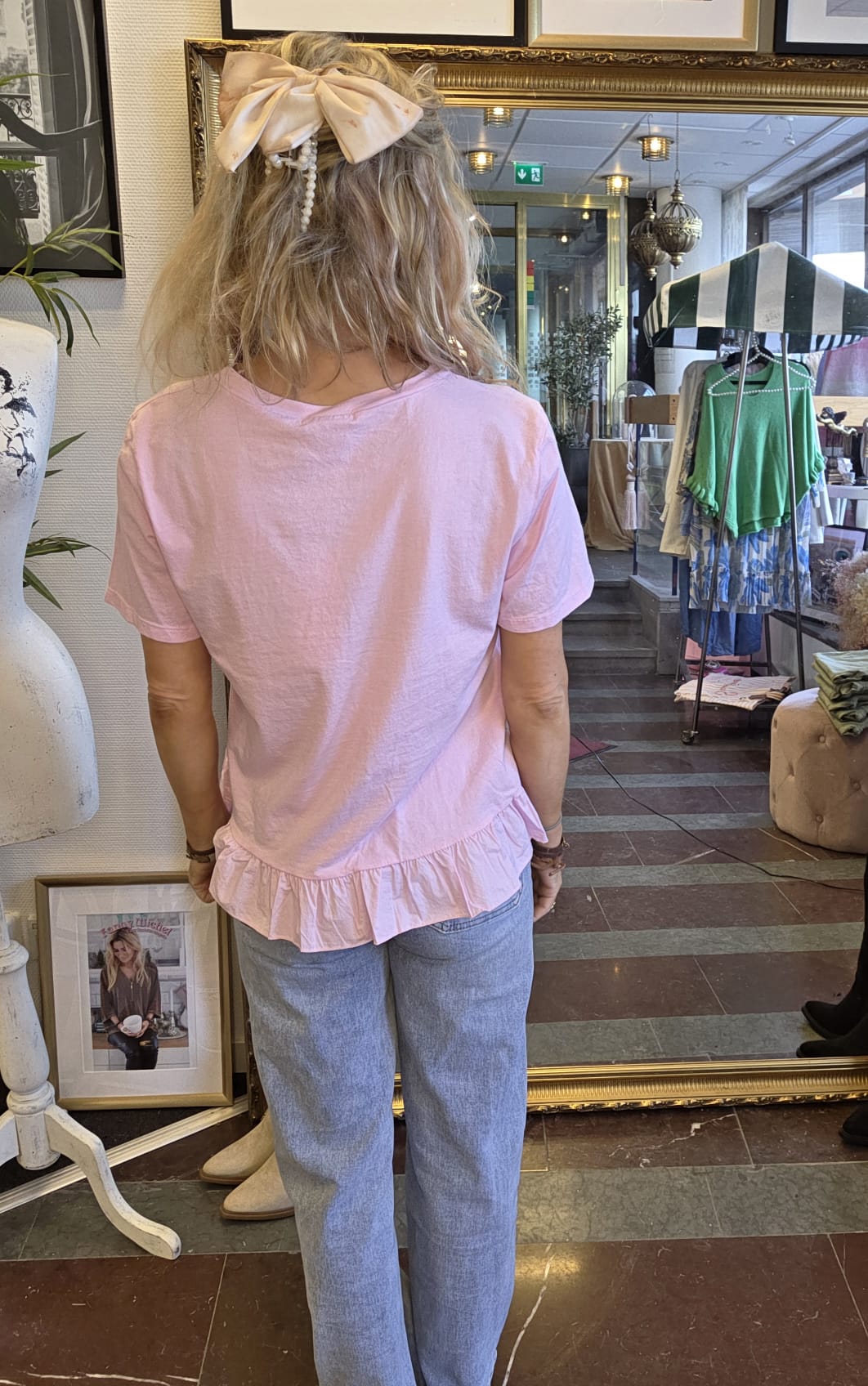 Kvinna sedd bakifrån står framför en stor guldspegel i en butik och visar sin outfit: ljusrosa topp med volangkant och ljusblå jeans, håret uppsatt med stor rosett. Klädställning syns i spegeln.