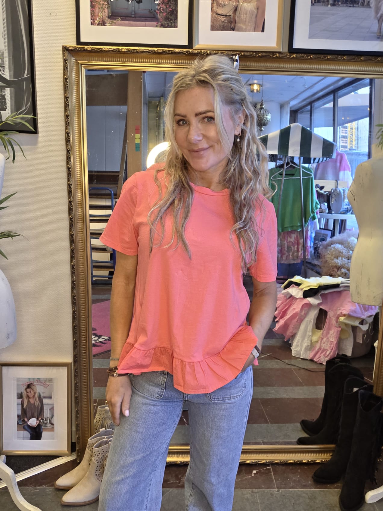 Leende kvinna med långt blont hår poserar framför en stor guldramsspegel i en klädbutik. Hon bär en korallrosa topp med volangkant och ljusa jeans, med handen i fickan; klädställningar och skor syns bakom.