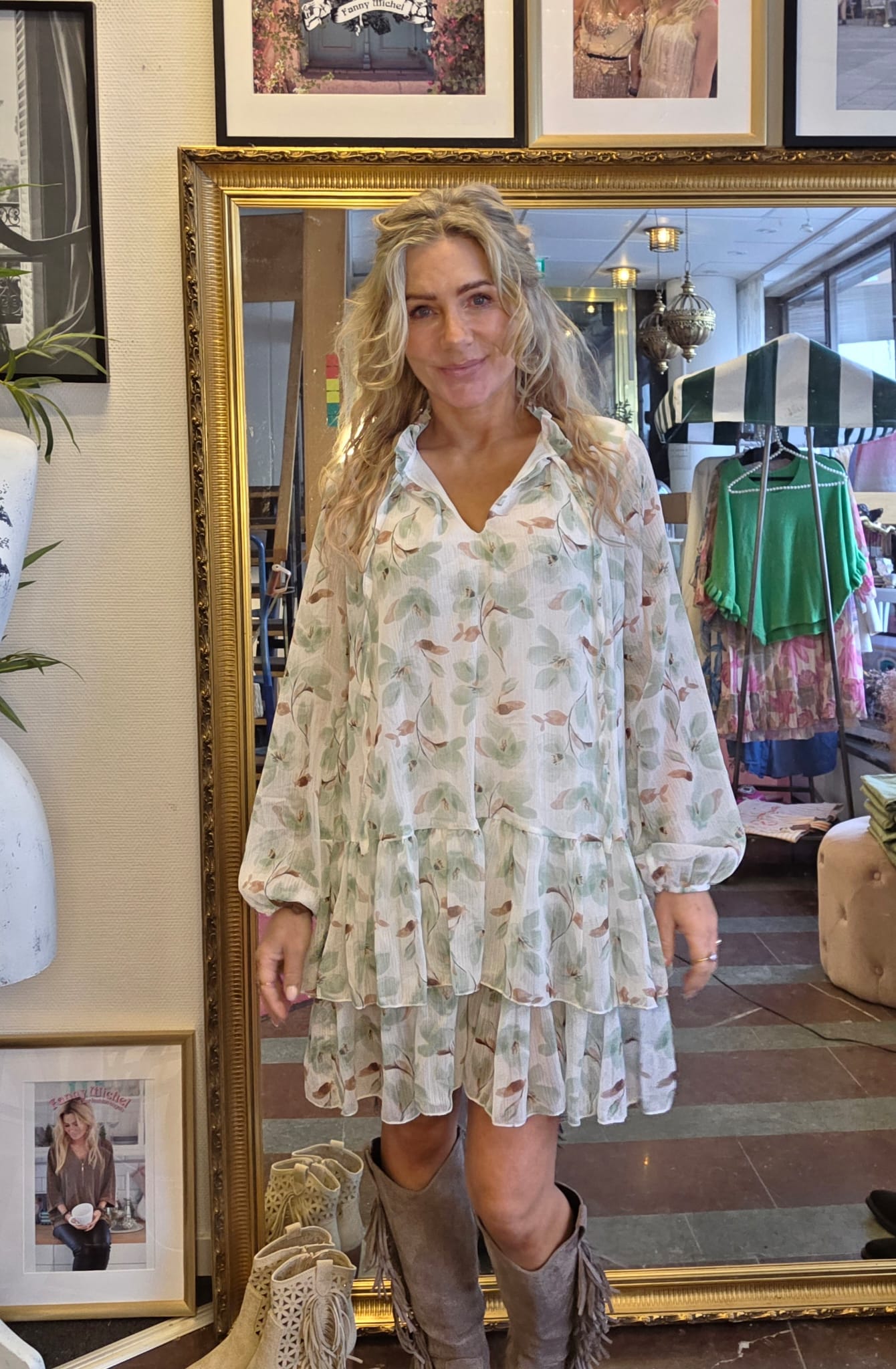 Kvinna står och ler framför en stor guldramsspegel i en klädbutik, iklädd ljus blommig volangklänning och fransboots. I bakgrunden syns klädställningar, randigt parasoll, lampor och inramade foton på väggen.