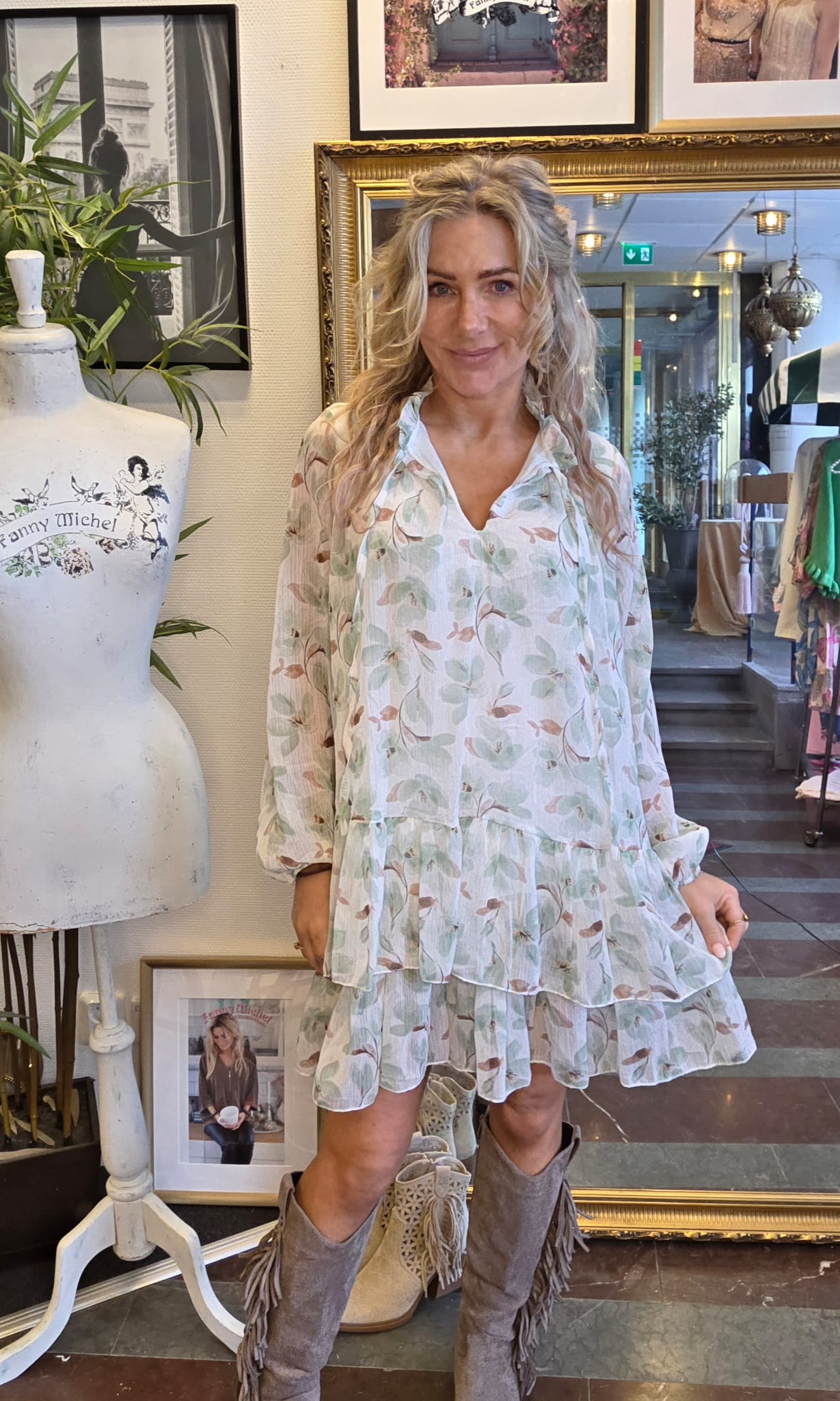 Kvinna står och ler i en klädbutik, iklädd ljus blommig volangklänning och fransstövlar. Hon håller ut kjolens kant. Bredvid syns provdocka med texten “Fanny Michel” samt inramade foton och spegel.