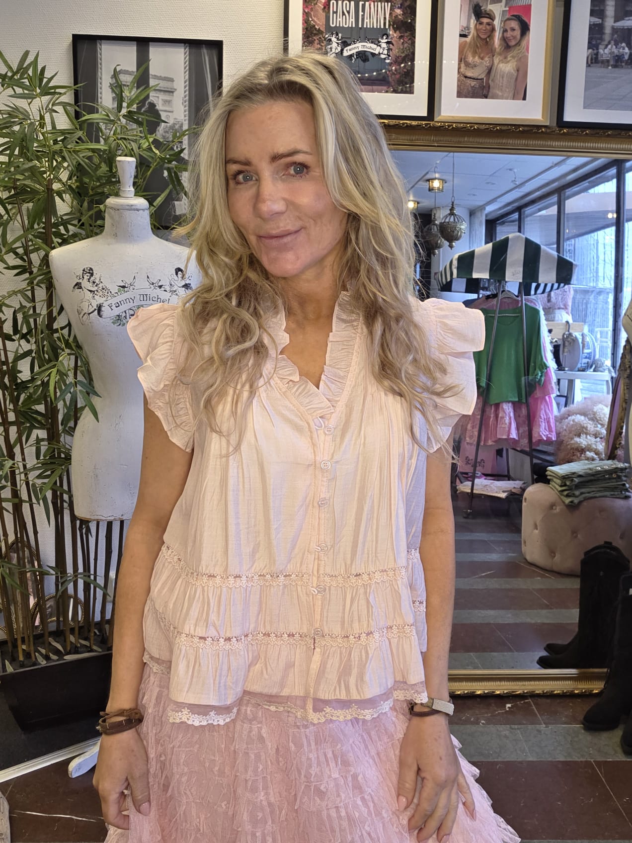 Blond kvinna står och ler mot kameran i en klädbutik, klädd i ljusrosa volangblus och kjol. Bredvid syns provdocka med texten “Fanny Michel” och på väggen en inramad skylt “CASA FANNY”.