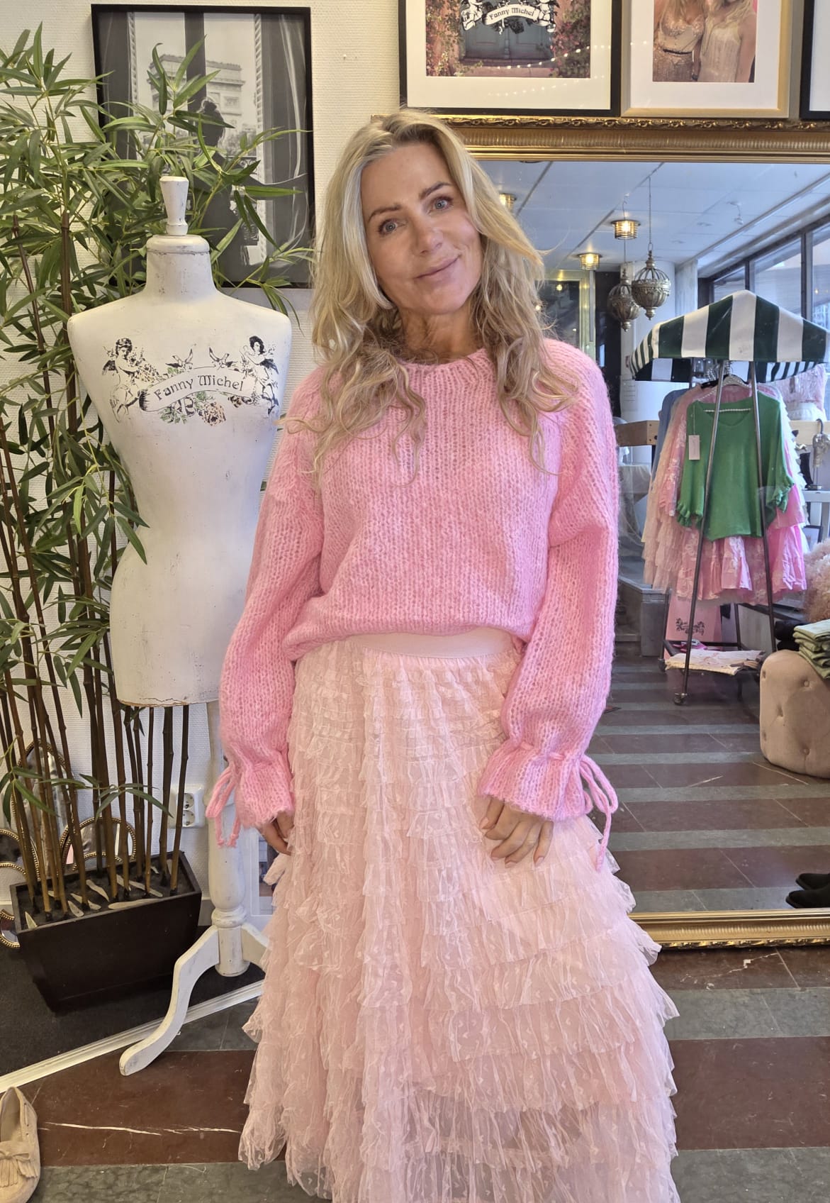 Blond kvinna står och ler i en klädbutik, iklädd rosa stickad tröja och lång, ljusrosa volangkjol. Bredvid står en provdocka med texten ”Fanny Michel”, och i bakgrunden syns klädställning och spegel.