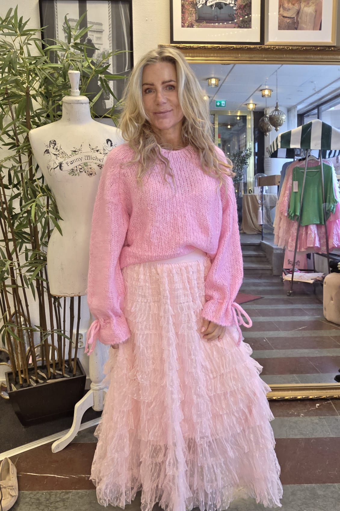 Blond kvinna står och poserar i en butiksmiljö, klädd i rosa stickad tröja och volangkjol i ljusrosa tyll. Bredvid syns en provdocka med texten ”Fanny Michel” samt klädställning och inredning i bakgrunden.