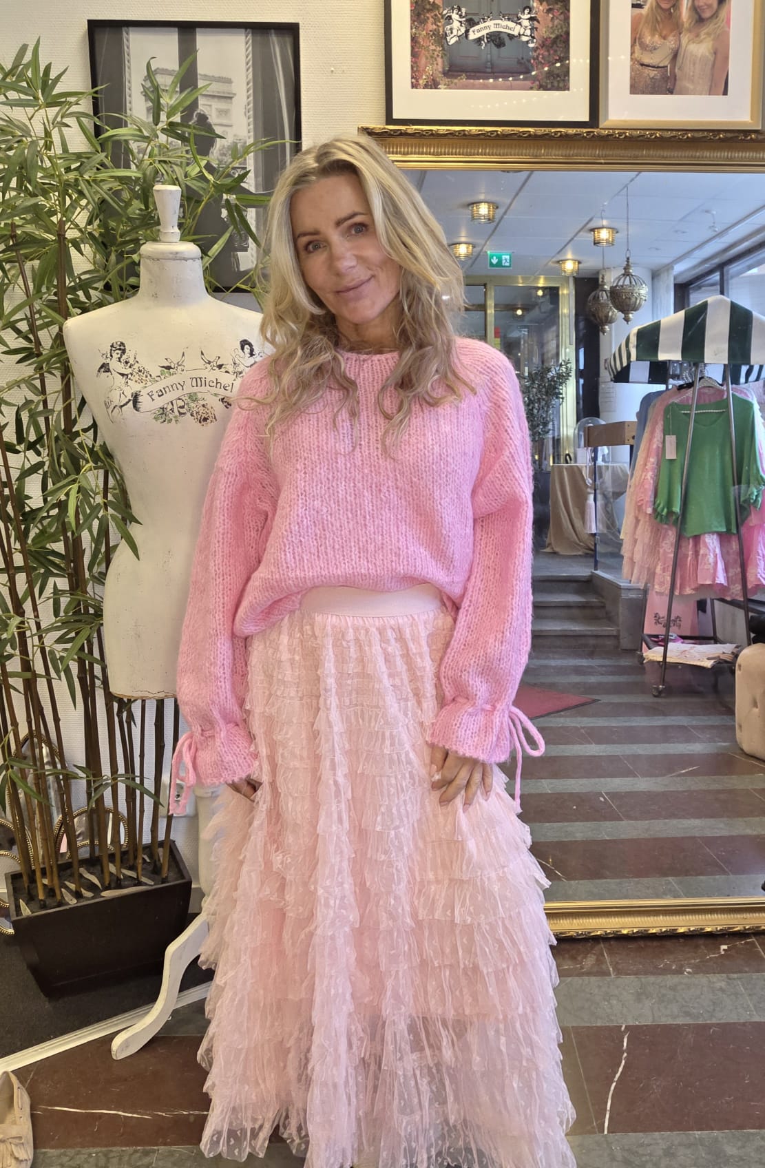 Blond kvinna ler mot kameran och poserar i en butiksmiljö, klädd i rosa stickad tröja och ljusrosa volangkjol. Bredvid står en provdocka med texten “Fanny Michel”, samt klädställning och spegel i bakgrunden.