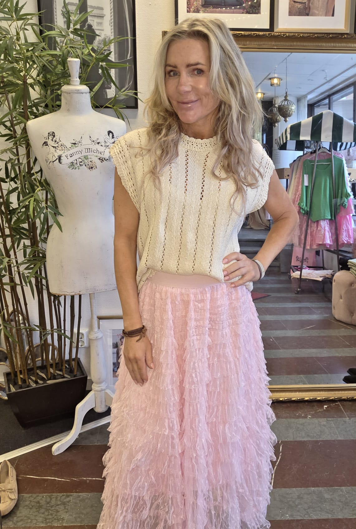 Kvinna med långt blont hår poserar med handen på höften i en butik, klädd i beige stickad topp och rosa volangkjol. Bredvid står en provdocka med texten “Fanny Michel”, och klädställningar syns i bakgrunden.