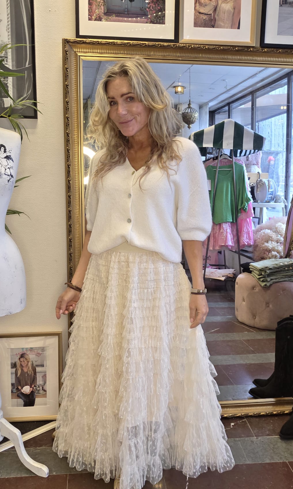 Kvinna med långt blont hår står och poserar framför en stor guldramsspegel i en butik, klädd i vit kofta och lång, skir volangkjol. I bakgrunden syns klädställning, fönster och dekor.