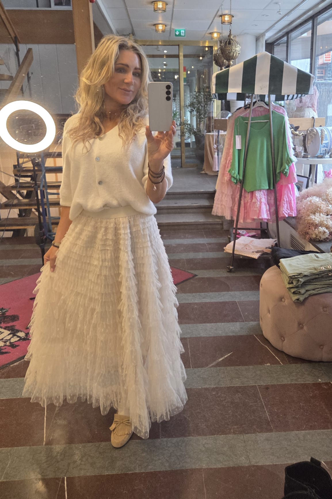 Blond kvinna tar en spegelselfie med mobil i en klädbutik, klädd i vit tröja och lång, volangig tyllkjol. Runt henne syns ringljus, klädställ med grönt och rosa plagg samt butikens entré.
