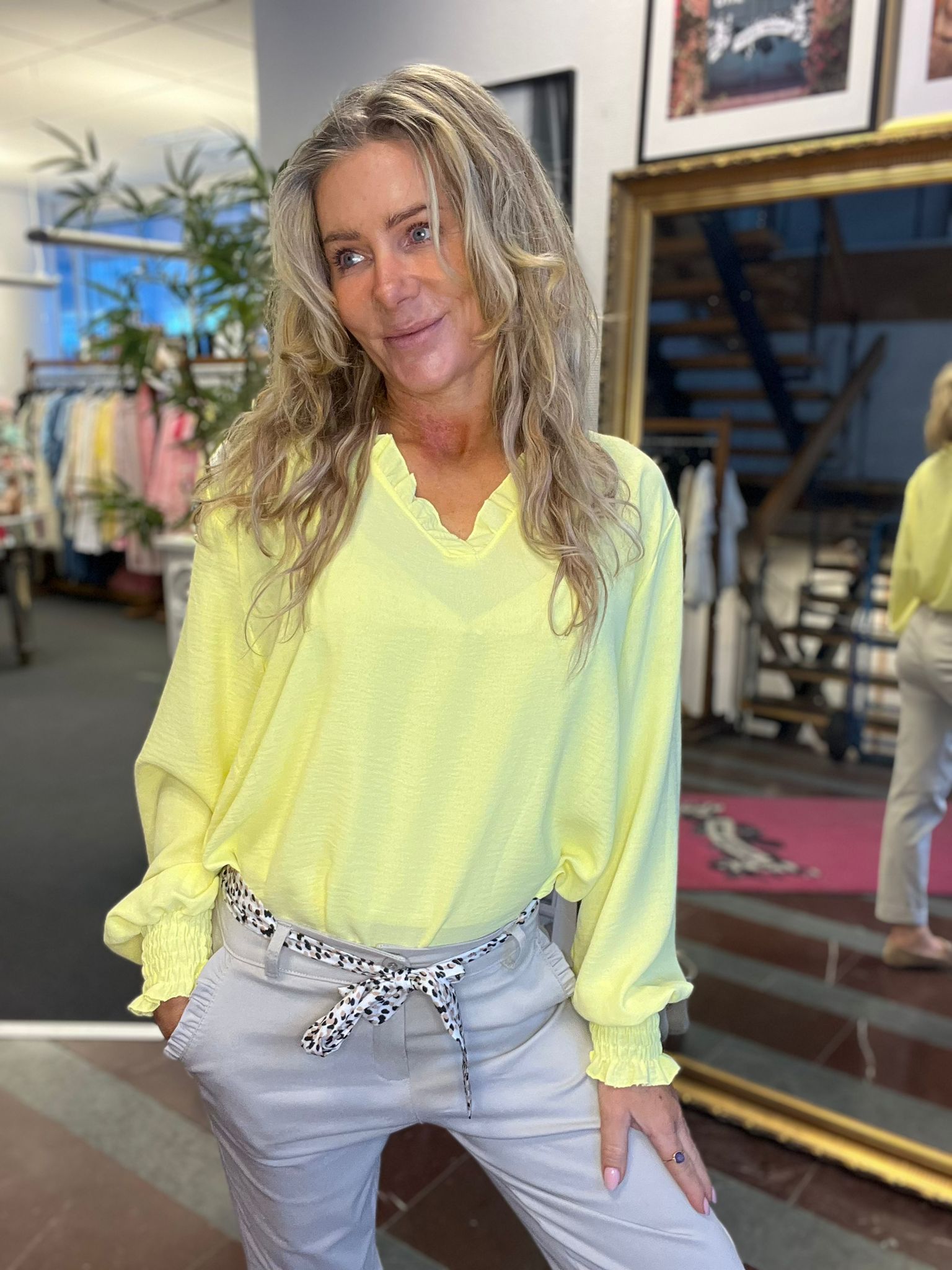 Kvinna med långt blont hår poserar och ler i en klädbutik, iförd limegul blus med volangkrage och ljusgrå byxor med mönstrat skärp. I bakgrunden syns klädställning, växter och en spegel.