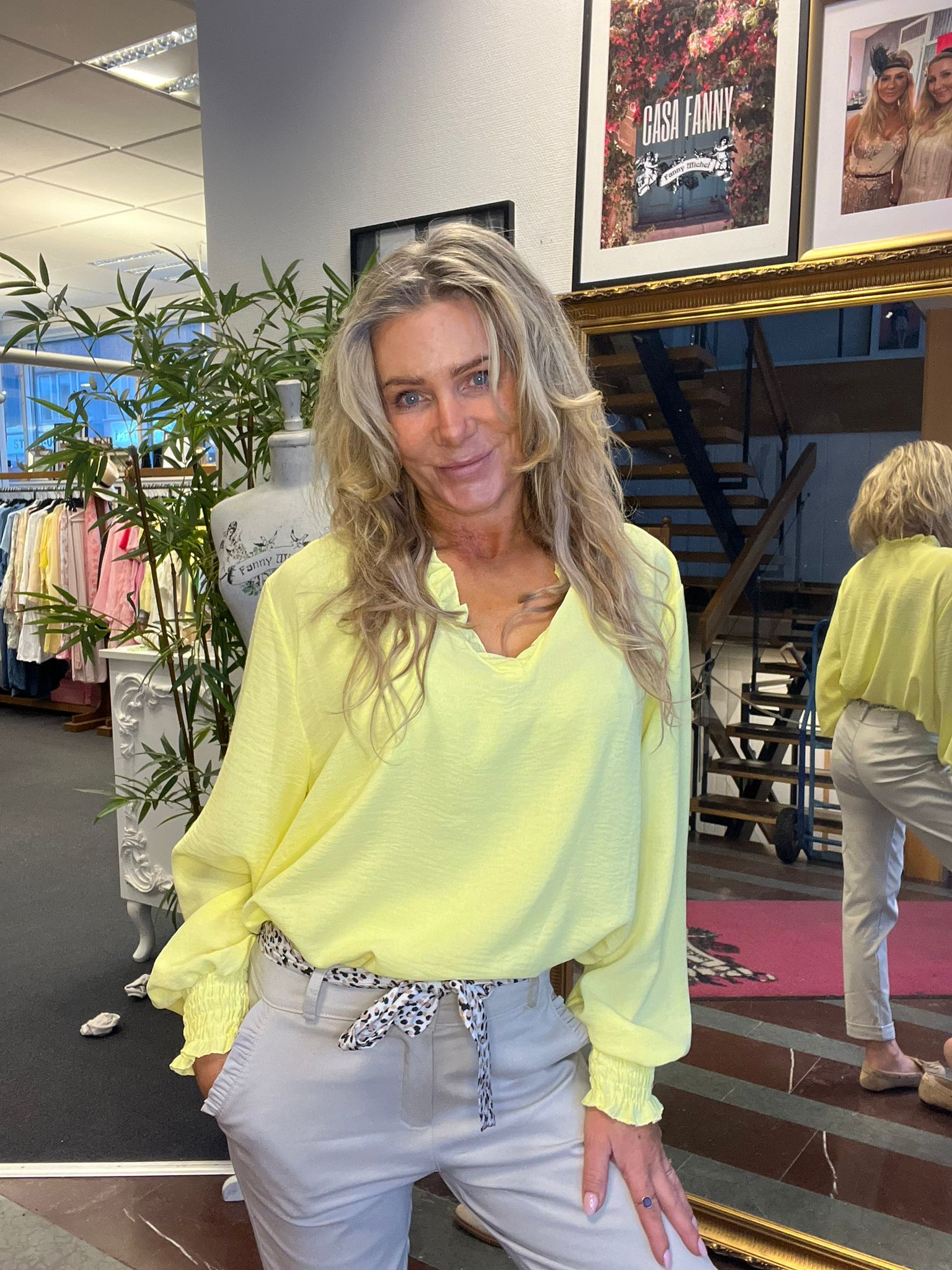 Kvinna i ljusgul blus poserar och ler lätt i en klädbutik, med handen i fickan. Bakom syns klädställ, spegel med hennes rygg och en tavla med texten “CASA FANNY”.