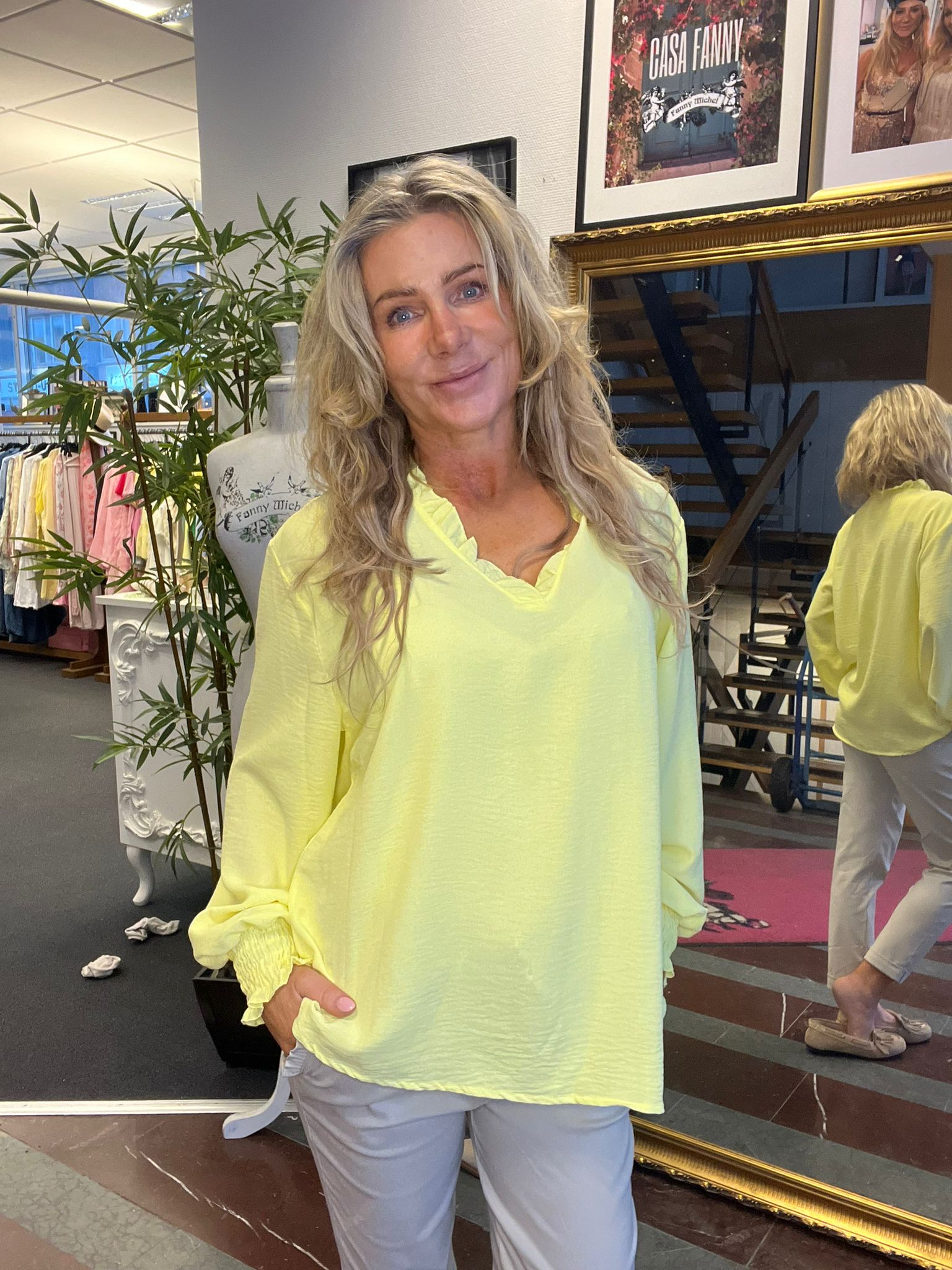 Kvinna med långt blont hår poserar och ler i en ljusgul blus och ljusa byxor i en klädbutik. Bakom syns klädställning, spegel och trappa samt en inramad skylt med texten “CASA FANNY”.