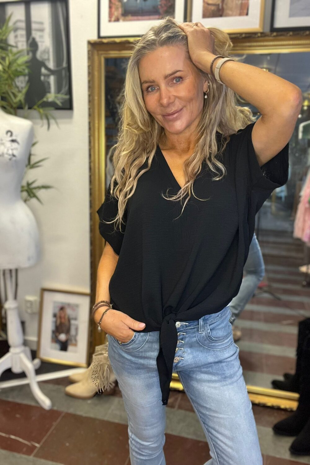 Blond kvinna i svart knuten blus och ljusa jeans poserar och ler, med ena handen i fickan och den andra i håret. Hon står i en inomhusmiljö med ramar på väggen, spegel och en provdocka.