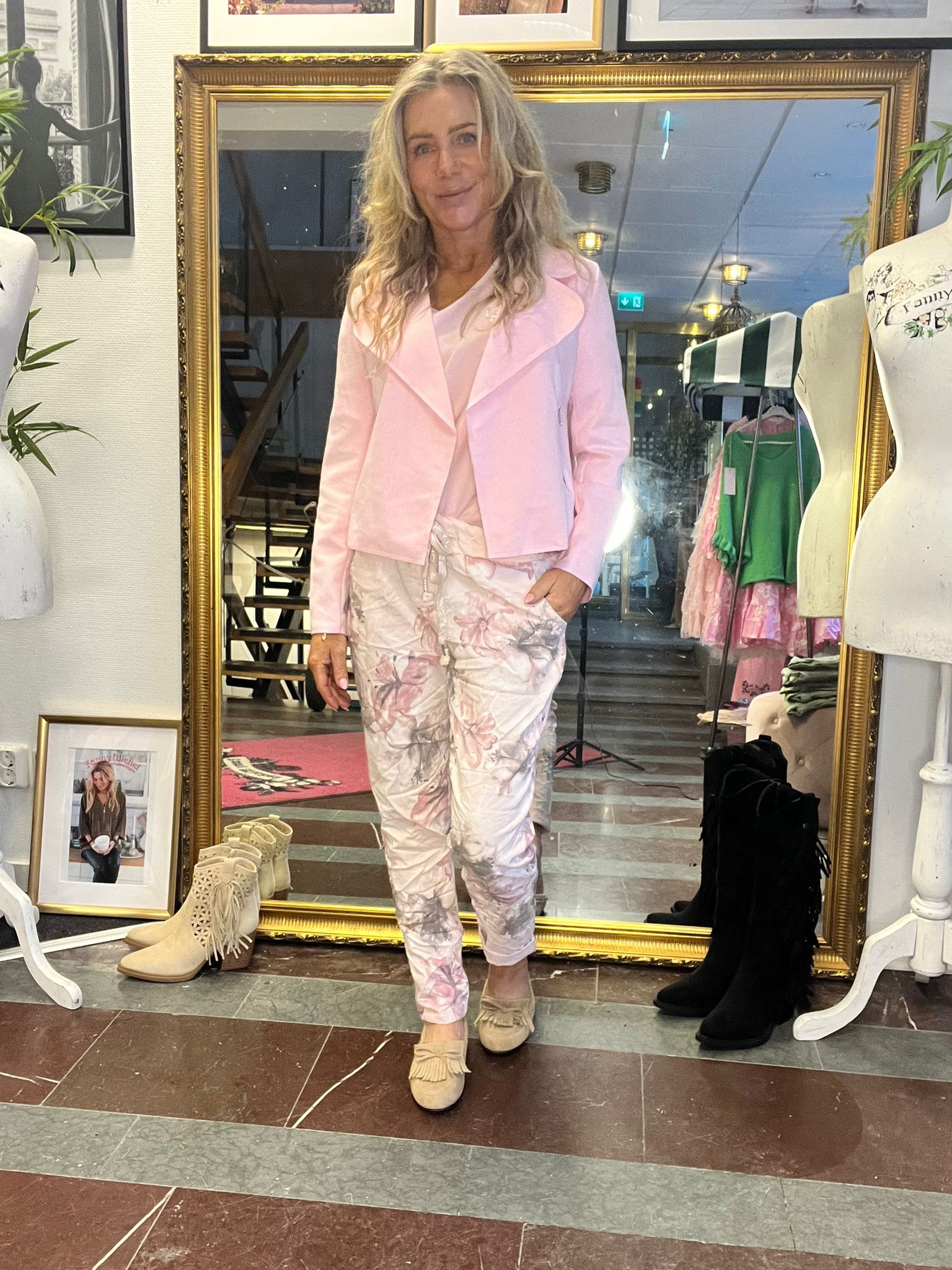 Blondi nainen poseeraa suuren kultakehyksisen peilin edessä, käsi taskussa, vaaleanpunaisessa jakussa ja vaaleissa kukkakuvioisissa housuissa. Ympärillä vaateboutique: mallinuket, ripustettuja vaatteita ja kenkiä lattialla.