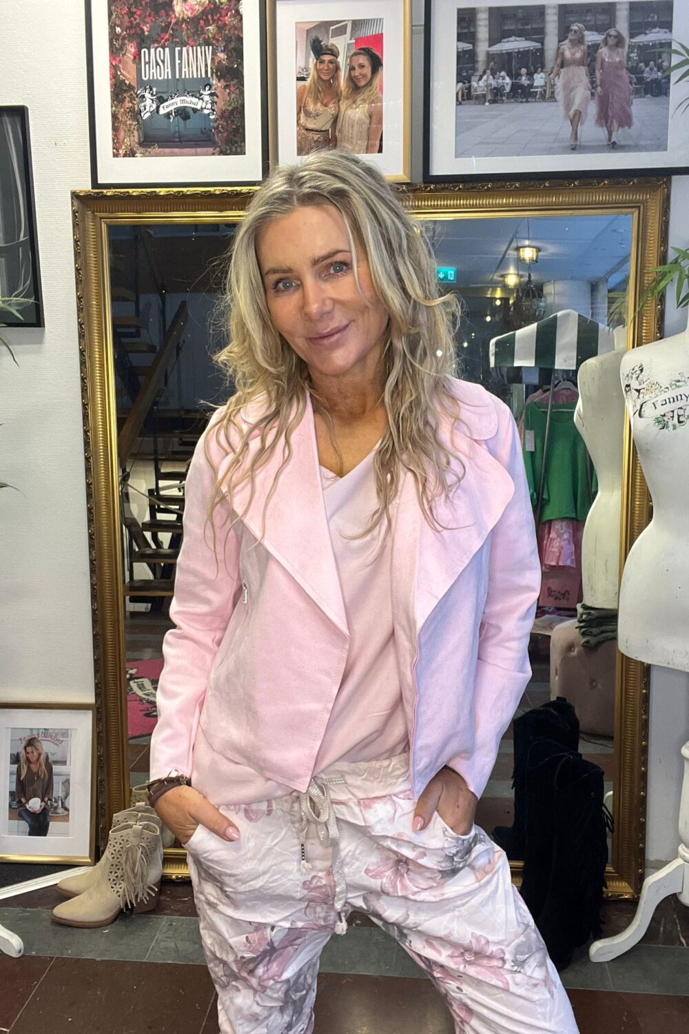 Leende kvinna i ljusrosa jacka poserar med händerna i fickorna framför en stor guldramsspegel i en butik. På väggen syns inramade foton och texten ”CASH FANNY” samt ”Fanny Michel”.