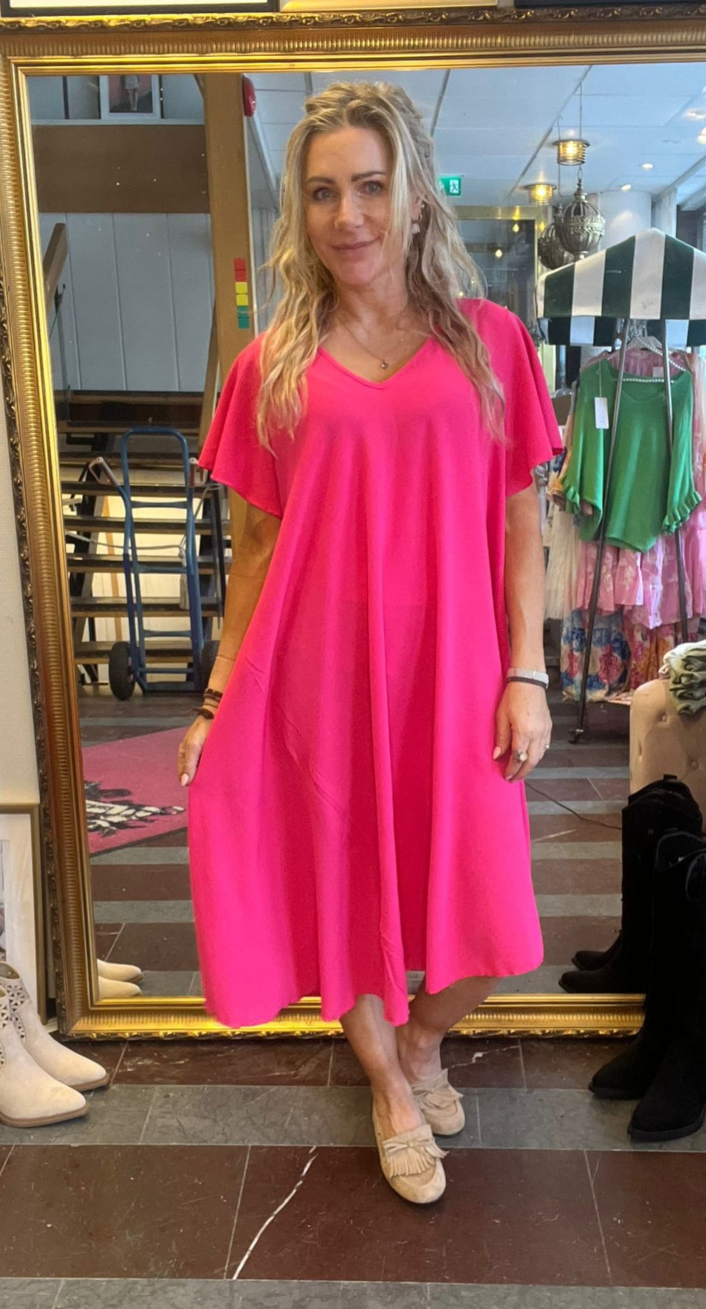 Kvinna står framför en stor guldramsspegel och visar upp en knallrosa, vid klänning med korta ärmar, matchad med beige loafers. Hon är i en butik med klädställningar och lampor i bakgrunden.