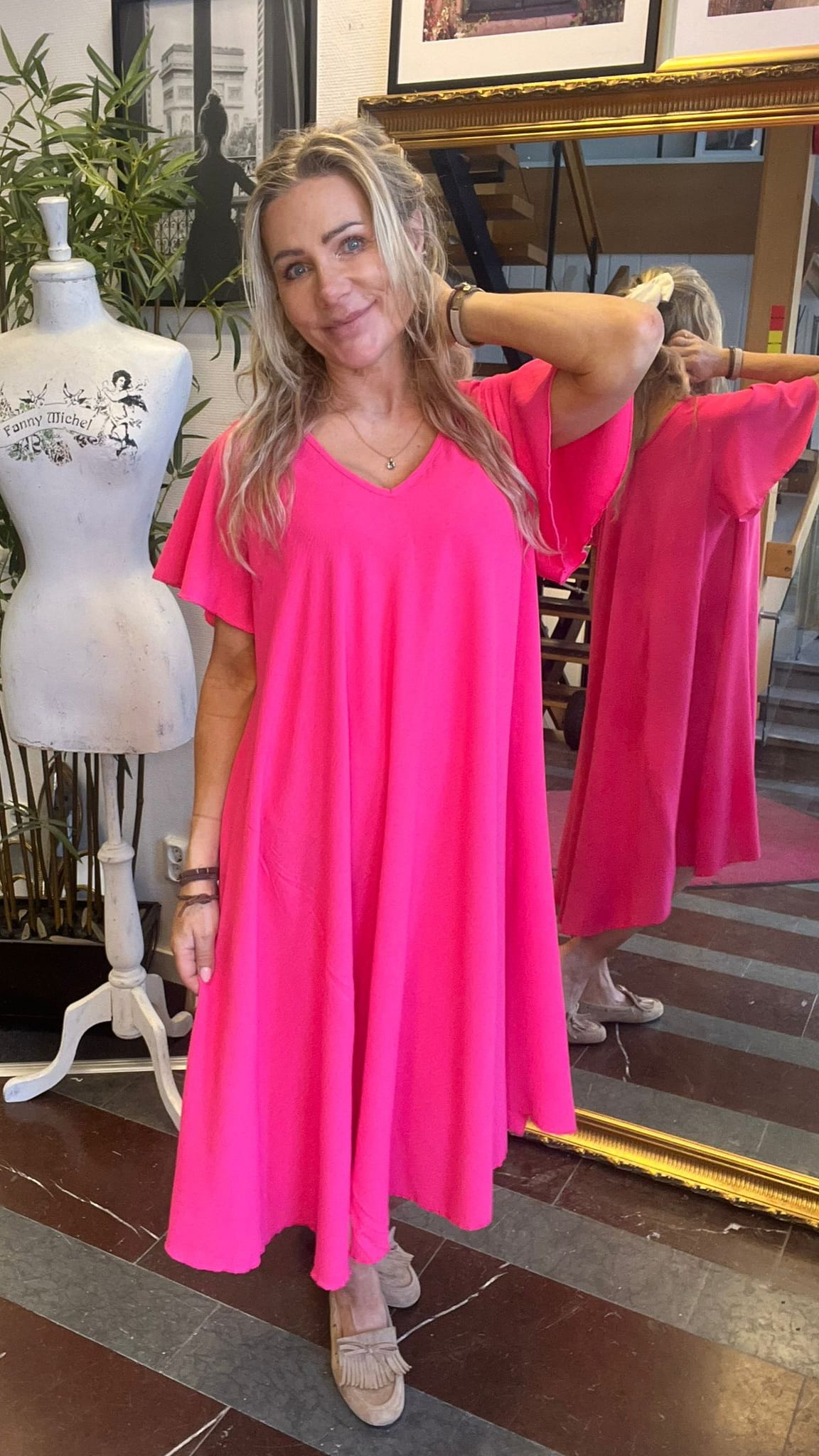 Kvinna i starkt rosa, löst sittande klänning poserar leende med handen bakom nacken framför en stor spegel i en butik eller studio. Bredvid står en provdocka med texten “Fanny Michel”.