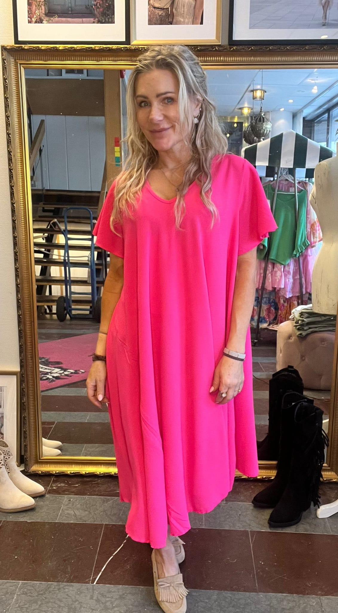 Kvinna med långt blont hår står och ler framför en stor guldramsspegel i en klädbutik, klädd i en starkt rosa, ledig klänning med fickor och beige loafers; klädställning och skyltdocka syns i bakgrunden.