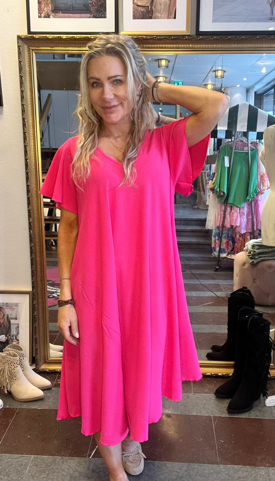 Kvinna i knallrosa, vid midiklänning poserar med ena handen bakom huvudet och ler mot kameran. Hon står i en butik framför en stor guldramsspegel, med kläder på ställning och boots i bakgrunden.