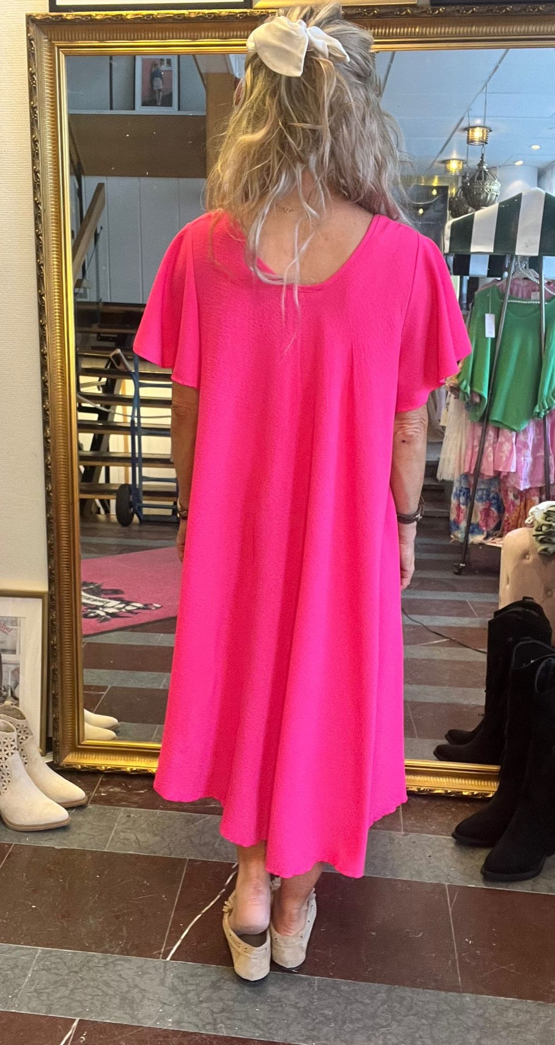 Kvinna står med ryggen mot kameran framför en stor guldramsspegel och provar en ceriserosa, vadlång klänning med korta ärmar. Håret är uppsatt med stor vit rosett; butiksmiljö med klädställ och skor.