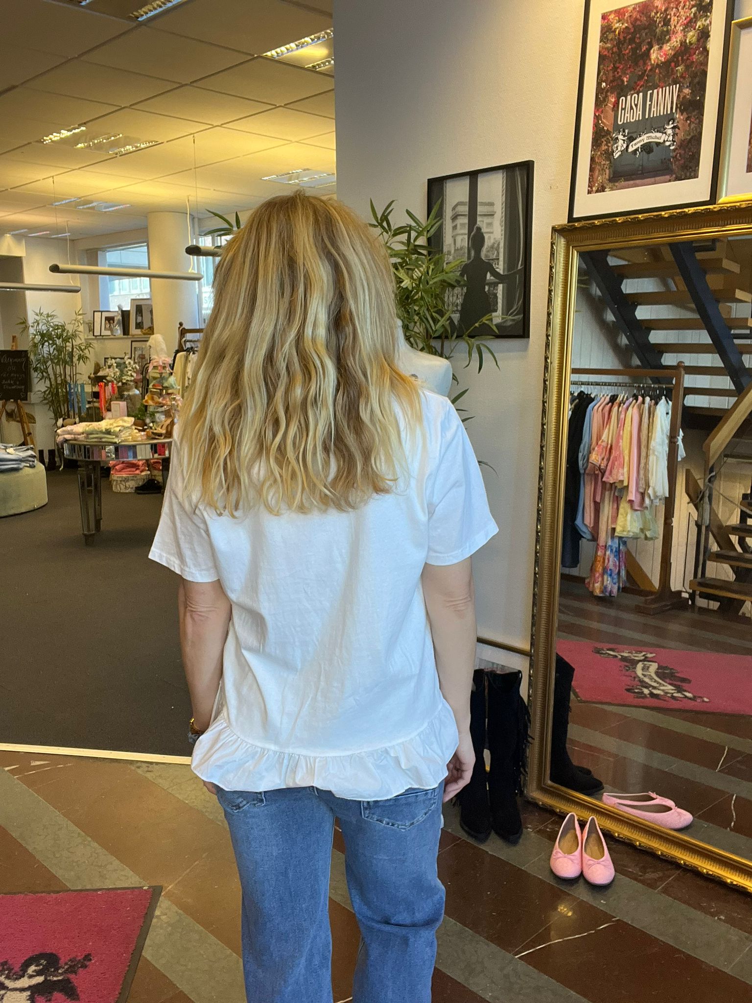 Kvinna med långt blont hår står med ryggen mot kameran och tittar mot en stor guldramsspegel i en klädbutik. Runt henne syns klädställningar, skor på golvet och en affisch med texten ”CASA FANNY”.
