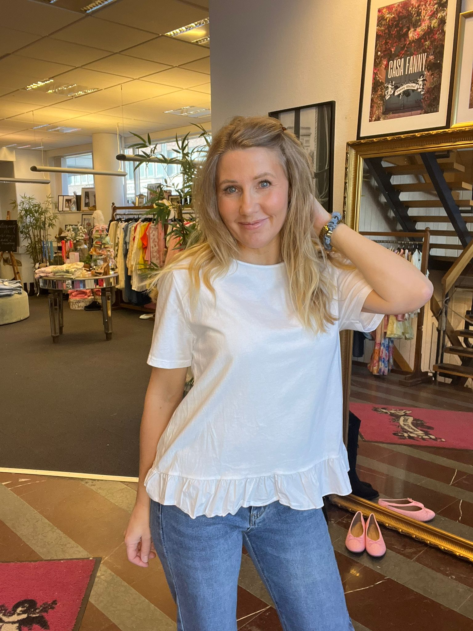 Leende blond kvinna i vit topp och jeans står och rör vid håret med ena handen i en klädbutik, med klädställningar och inredning bakom. På väggen syns texten “CASA FANNY”.
