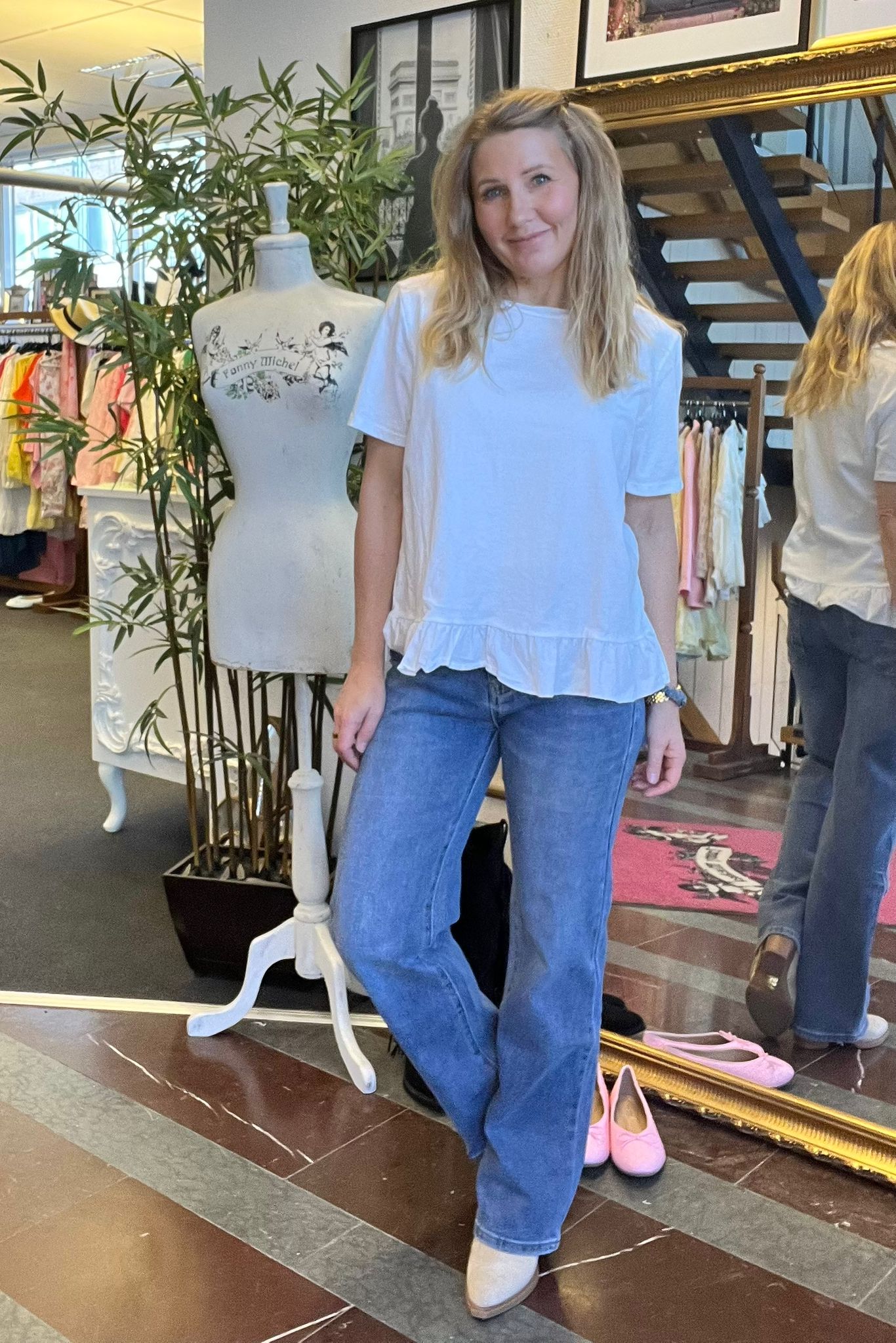 Leende kvinna poserar i en klädbutik, klädd i vit topp med volangkant och blå jeans. Hon står bredvid en provdocka med texten “Fanny Michel”, med klädställningar, växter och en spegel i bakgrunden.