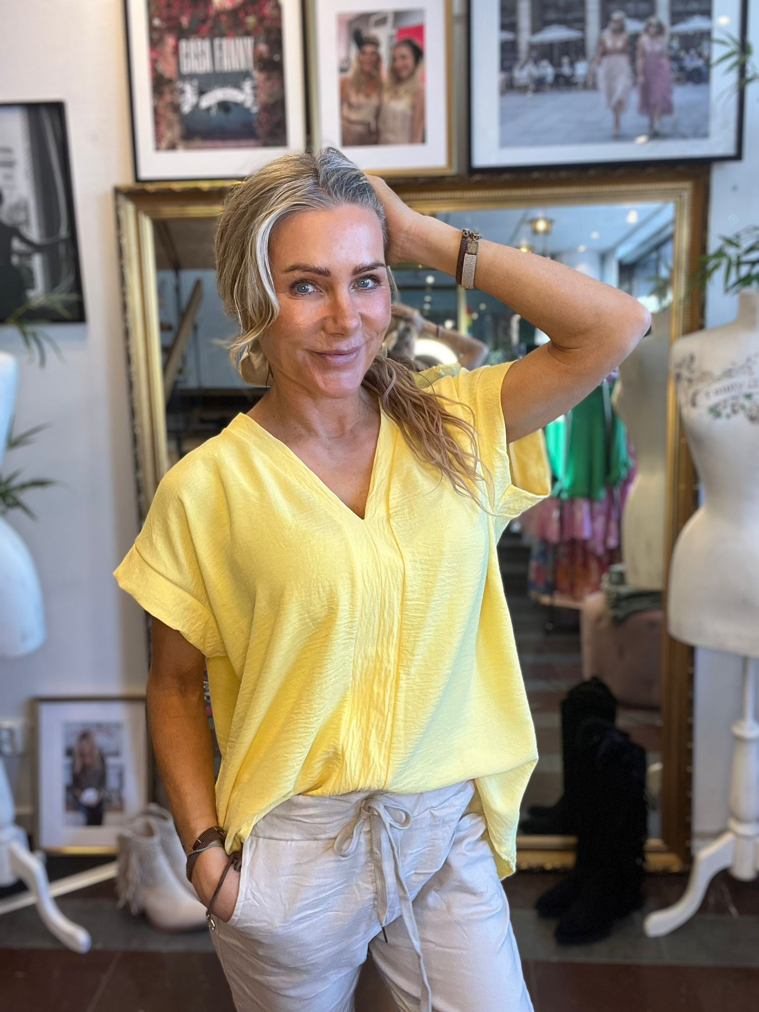 Leende kvinna i gul topp och ljusa byxor poserar med ena handen i håret och den andra i fickan. Hon står i en butik eller studio med stor spegel, provdocka och inramade foton på väggen.