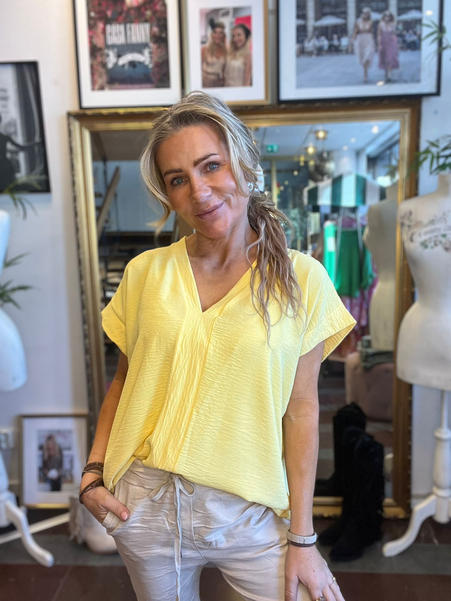 Leende kvinna i gul V-ringad topp och ljusa byxor poserar framför en stor guldramsspegel i en butik eller ateljé, med inramade foton på väggen och en provdocka bredvid. Text i bakgrunden: “GIRL BOSS”.