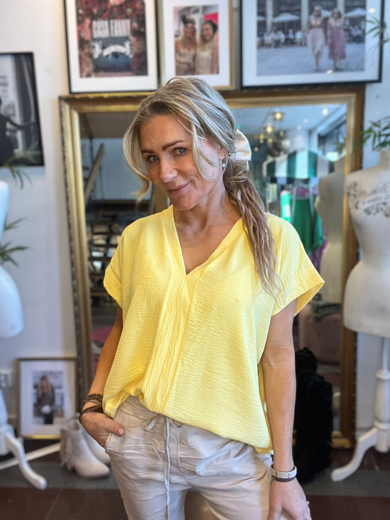 Blond kvinna ler mot kameran och posar med ena handen i fickan, klädd i gul blus och beige byxor. Hon står i en ateljé/butik med stor guldramsspegel, provdockor och inramade foton på väggen.