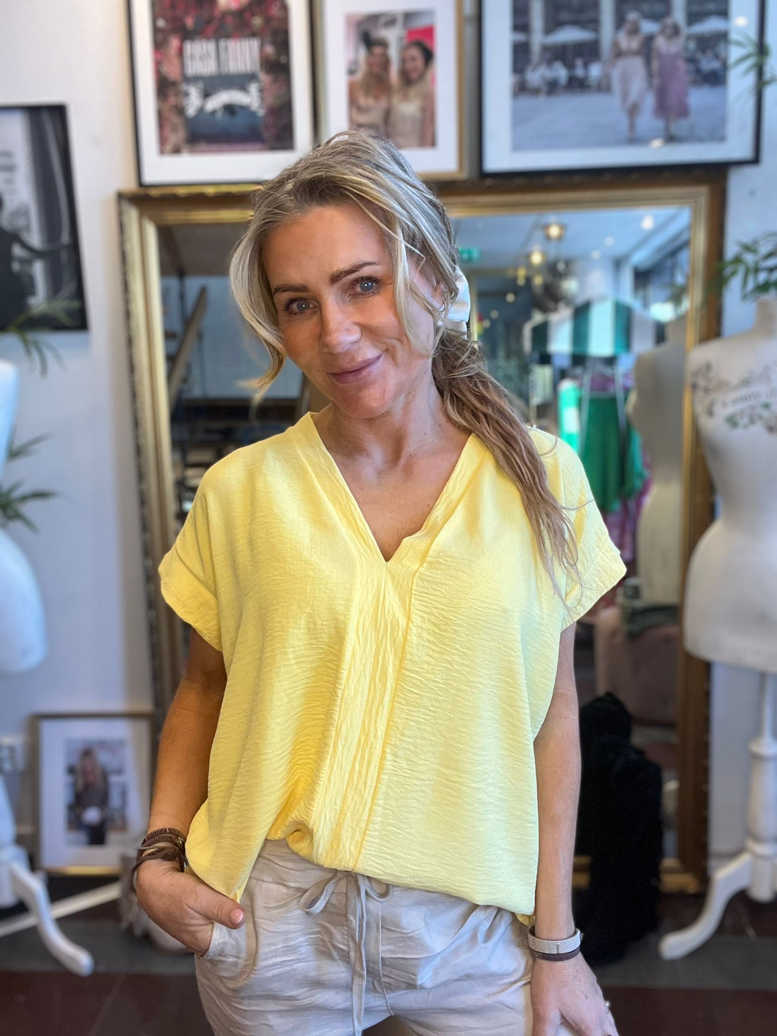 Blond kvinna ler mot kameran och står avslappnat med handen i fickan, klädd i gul topp och ljusa byxor. I bakgrunden syns en klädbutik med provdockor, spegel och inramade foton på väggen.