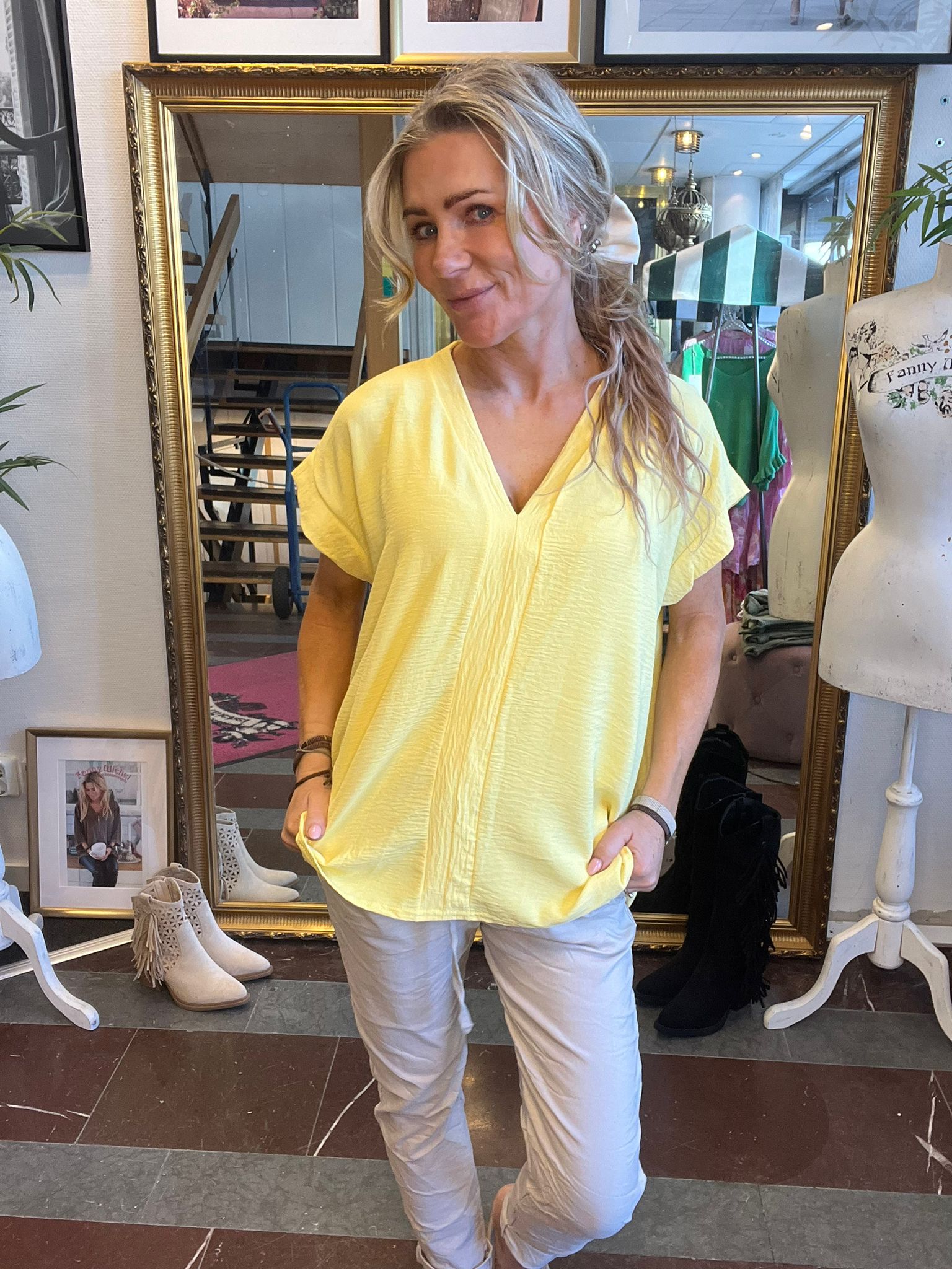 Leende kvinna med blont hår poserar framför en stor guldramsspegel och håller ut sin gula tunika. Hon bär ljusa byxor. I bakgrunden syns klädbutiksmiljö med skyltdocka, klädställning och skor. Text på skyltdocka: ”Fanny”.