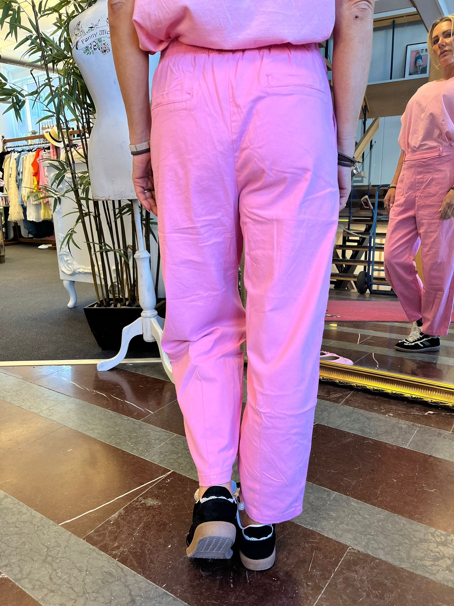 Person i rosa jumpsuit står med ryggen mot kameran framför en spegel i en klädbutik, som visar framifrån. Klädställning, bambuväxt och provdocka syns bredvid; text på provdockan: “Fanny mic”.