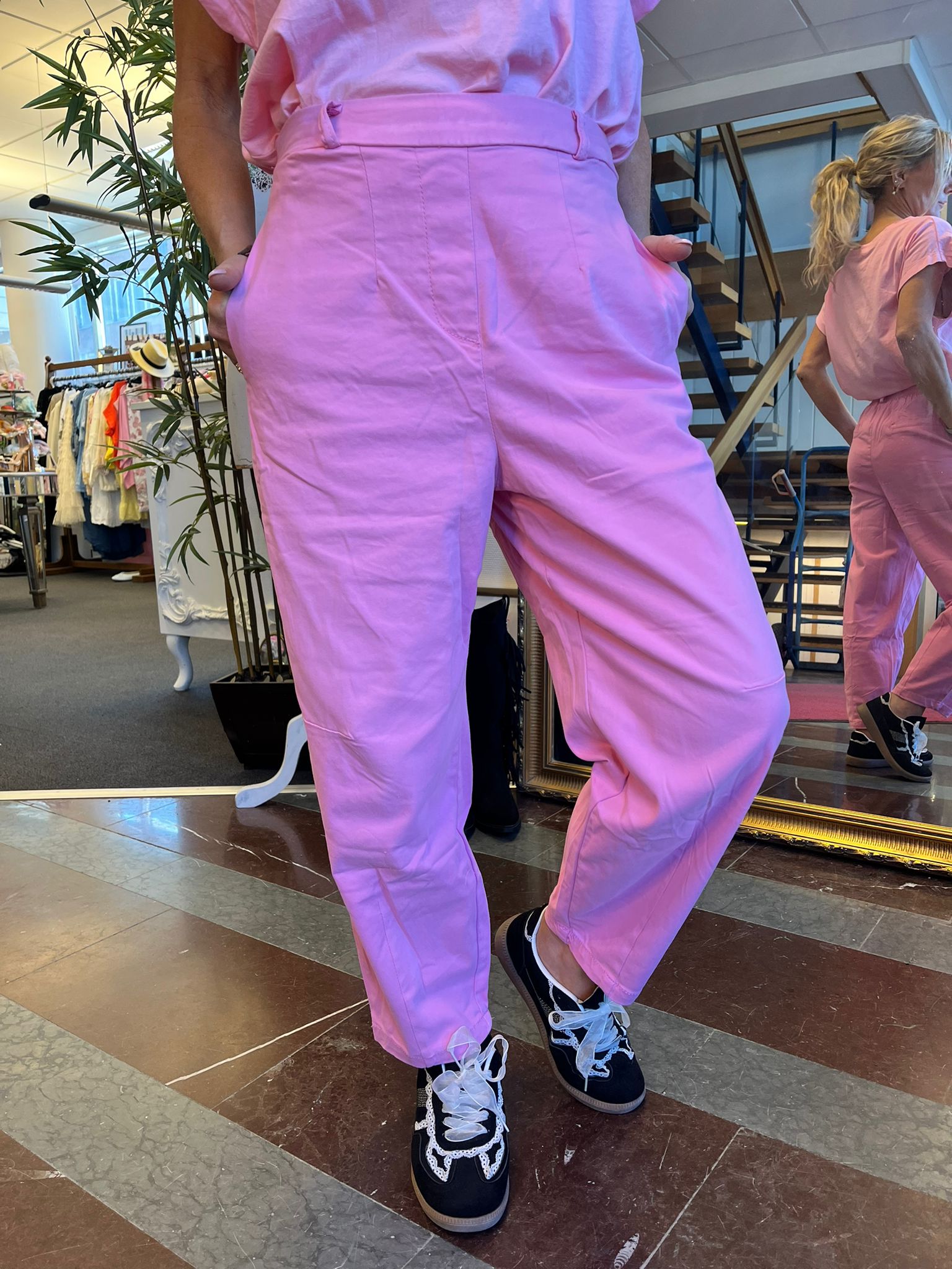 Person modellerar ljusrosa byxor med händerna i fickorna och ena benet lätt böjt, tillsammans med matchande topp och svarta sneakers med vita snören. Miljö: klädbutik med klädställning, spegel och trappa i bakgrunden.