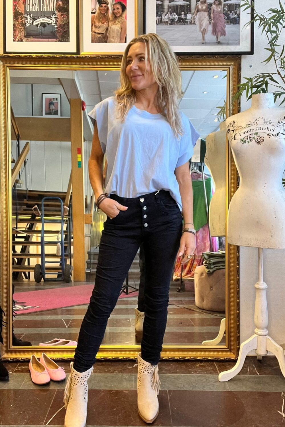 Blond kvinna står och poserar framför en stor guldramsspegel i en butik, med handen i fickan och blicken åt sidan. Bredvid syns en provdocka med texten “Fanny Michel” och väggbilder med “CASA FANNY”.
