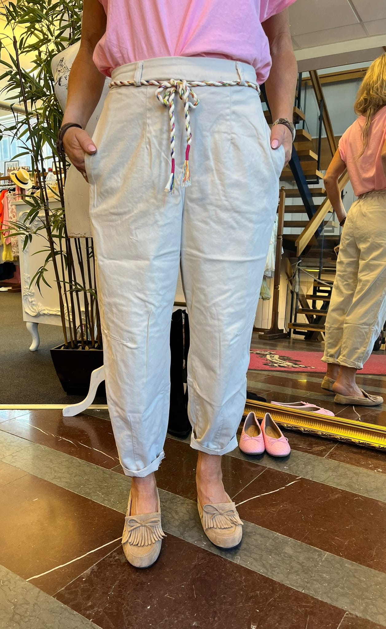 Person står i en klädbutik och visar vita, högmidjade byxor med sidfickor och uppvikta benslut, knutna med ett flätat flerfärgat repbälte; rosa topp och beige loafers syns, spegel och skor i bakgrunden.