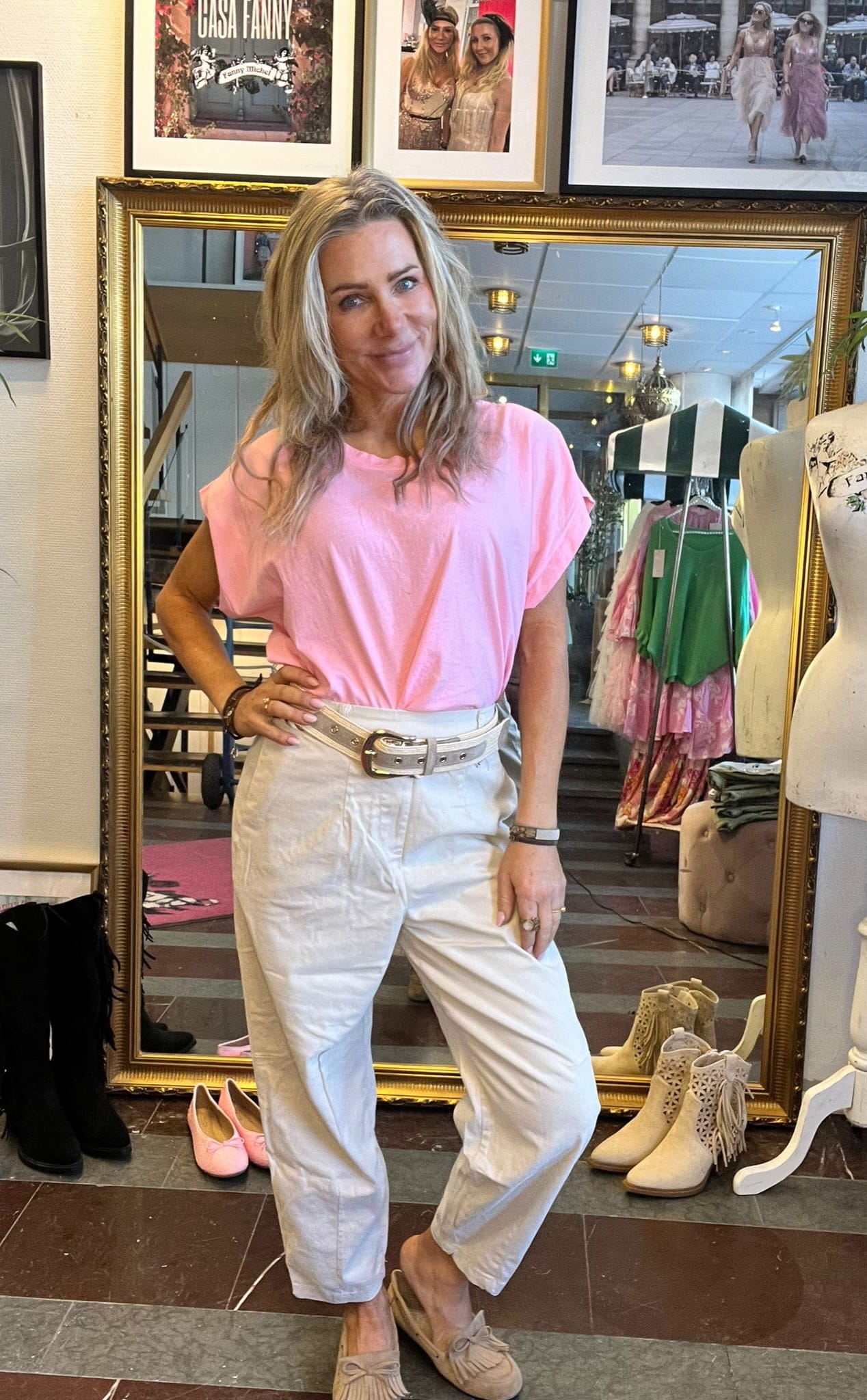 Kvinna med långt blont hår poserar med handen på höften framför en stor guldramad spegel i en klädbutik, klädd i rosa topp och vita byxor. Ovanför syns ramar med texten “CASA FANNY”.