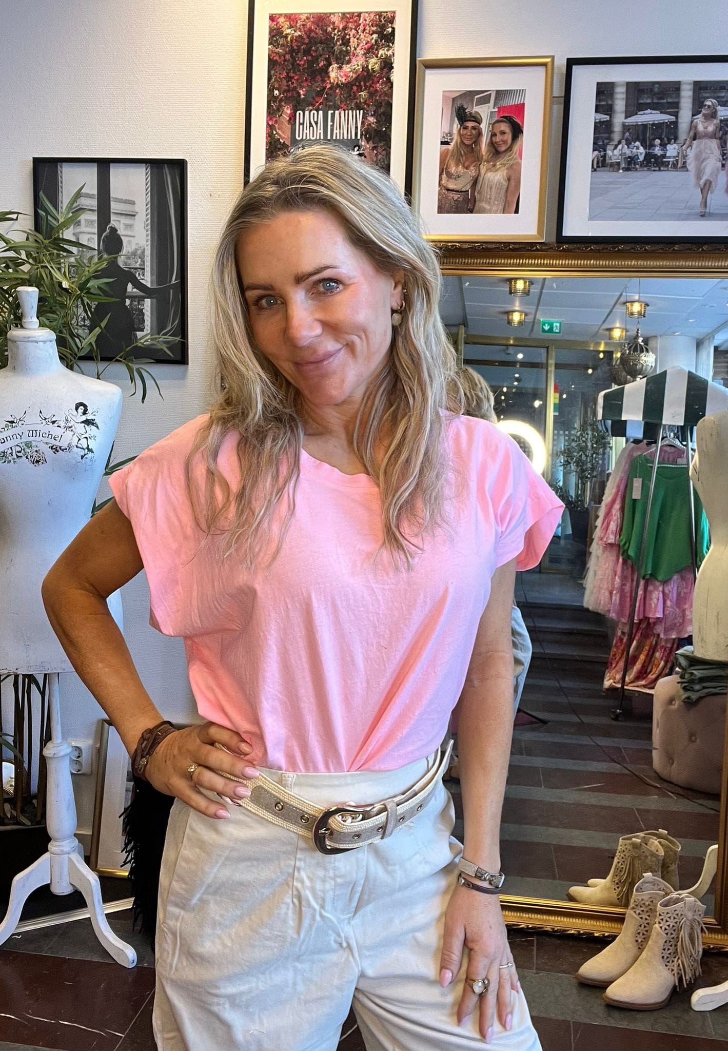 Kvinna med långt blont hår står och ler med handen på höften i en klädbutik, klädd i rosa topp och ljusa byxor. Bakom syns provdocka, klädställning och inramade foton med texten “CASA FANNY”.