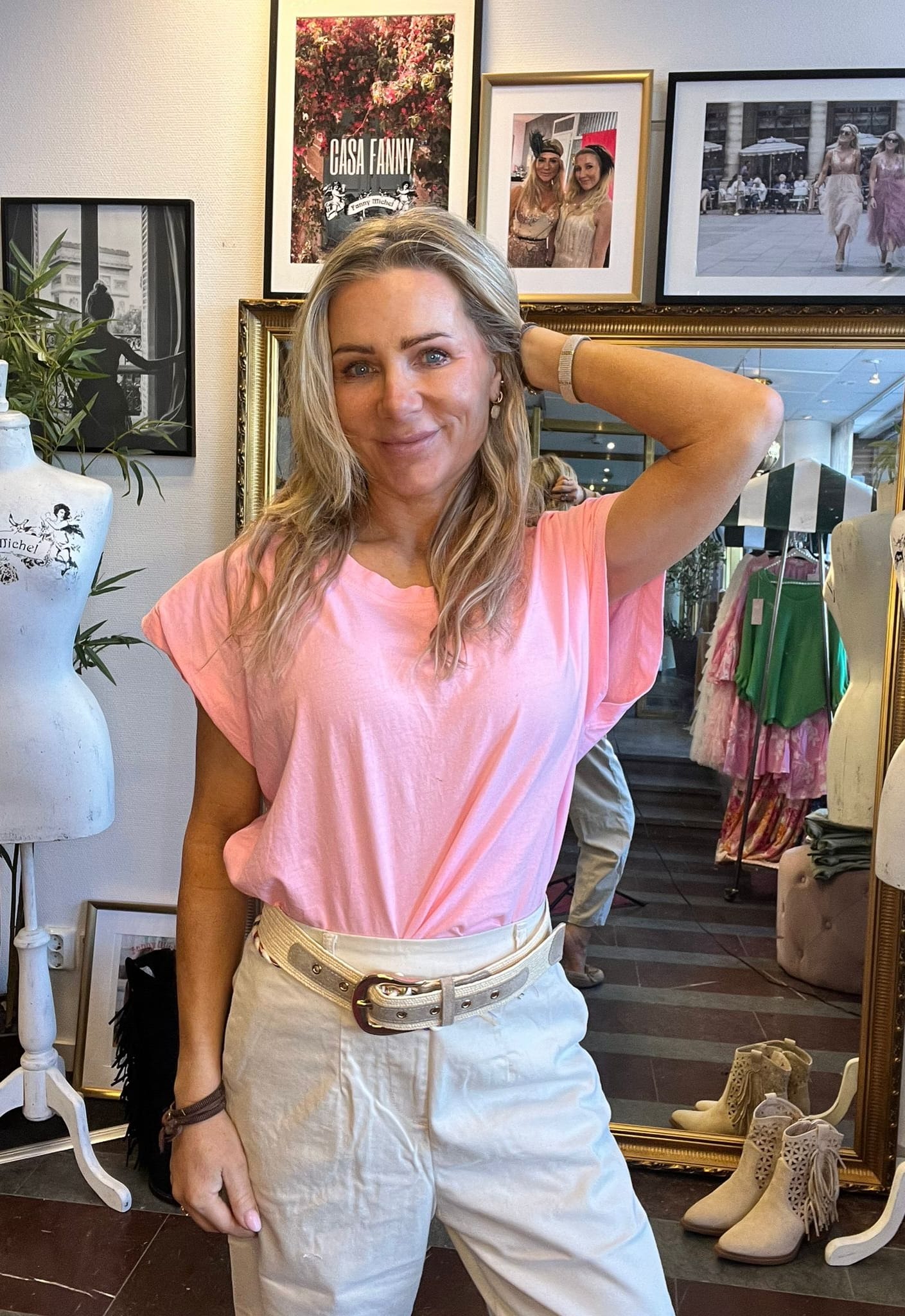 Kvinna i rosa t-shirt och vita byxor poserar leende med ena handen bakom huvudet i en klädbutik. Bakom henne syns spegel, klänningar, skyltdockor och inramade foton samt affisch med texten “CASA FANNY”.