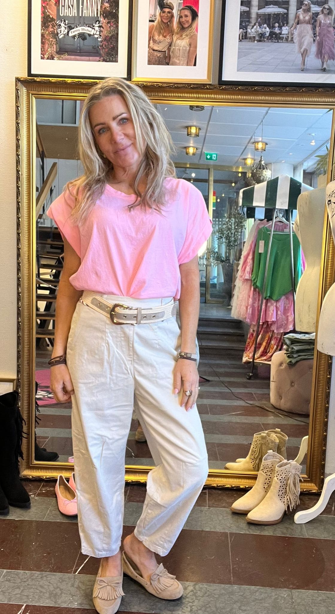Kvinna står och poserar framför en stor guldramsspegel i en klädbutik, klädd i rosa topp, vita byxor med brett bälte och beige loafers. I bakgrunden syns kläder på ställ och texten “CASA FANNY”.