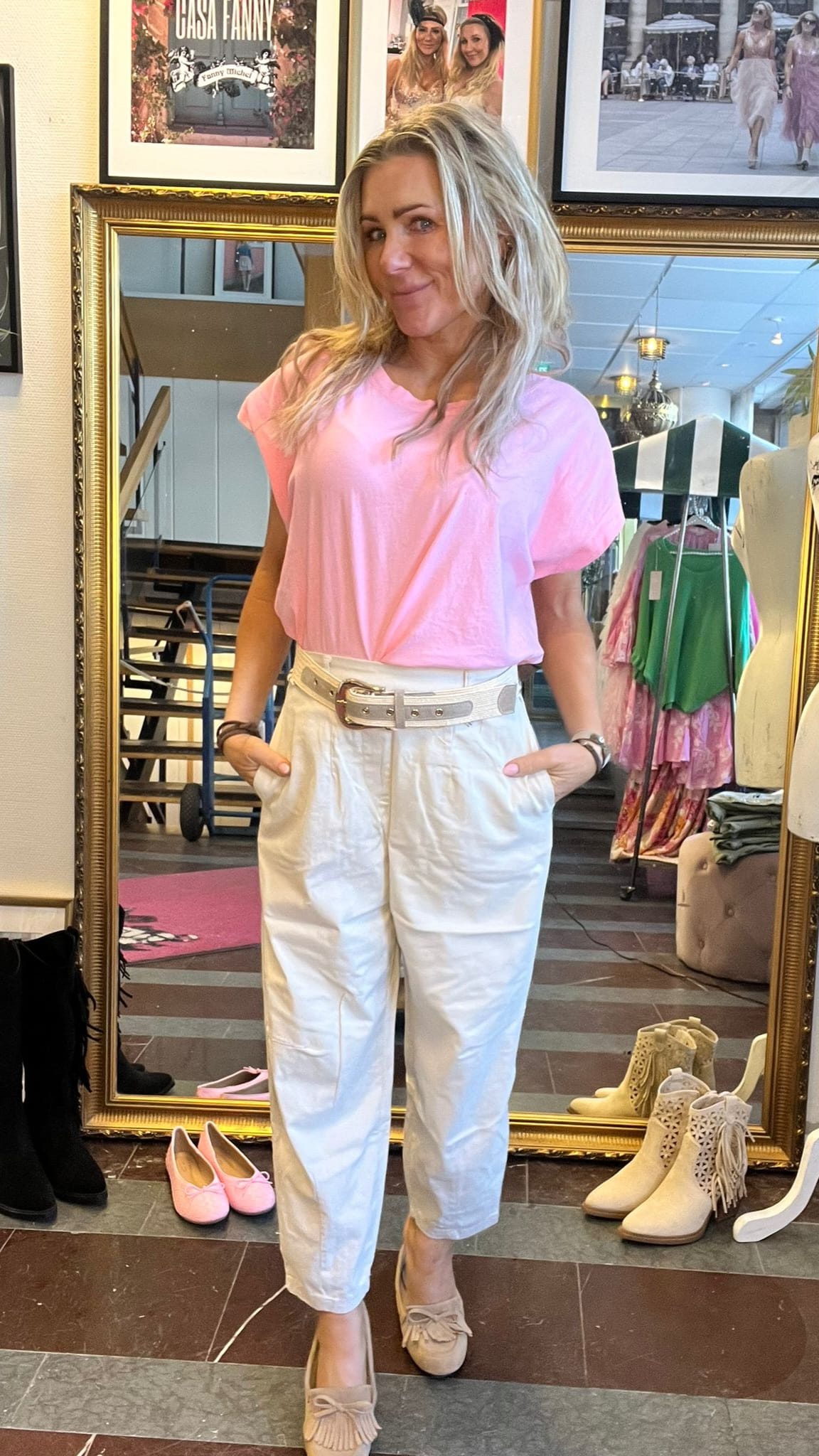 Blond kvinna poserar med händerna i fickorna framför en stor guldramsspegel i en klädbutik, klädd i rosa topp och vita byxor med bälte. Skor ligger på golvet; inramade foton ovanför, text: “CASA FANNY”.