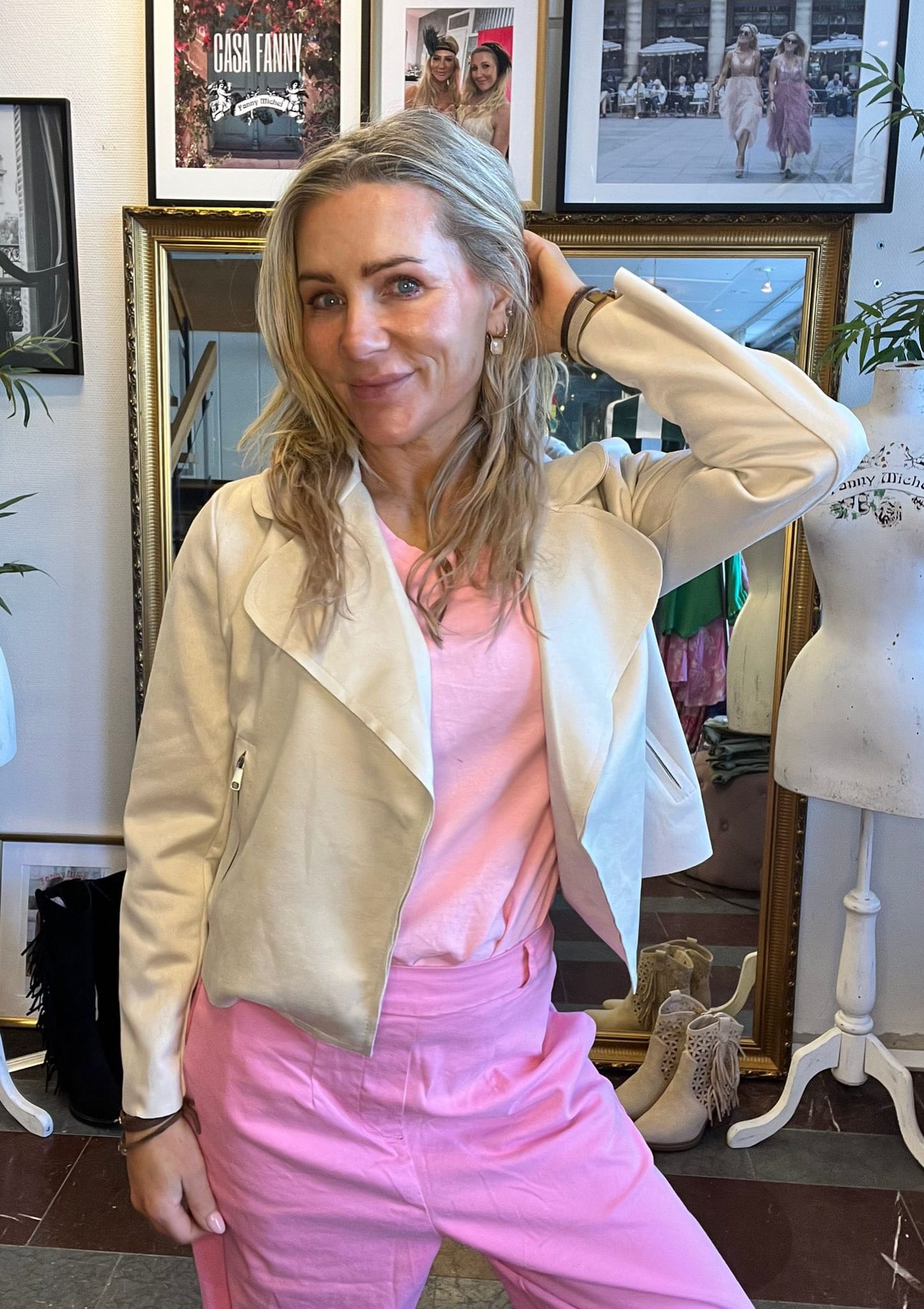 Leende kvinna med blondt hår poserar inomhus med ena handen bakom huvudet, klädd i ljus jacka och rosa outfit. Bakom henne syns speglar, en provdocka och inramade foton med texten “CASA FANNY”.