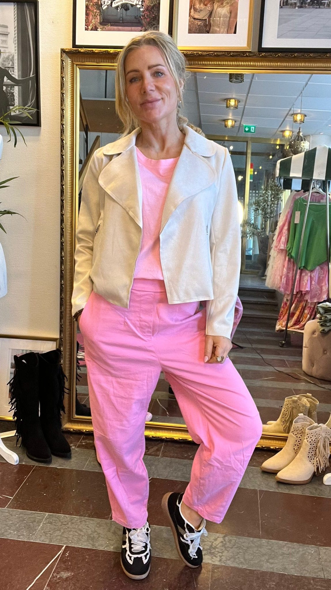 Blond kvinna poserar med ena handen i fickan framför en guldramsspegel i en butik, klädd i ljus jacka, rosa tröja och rosa byxor samt svartvita sneakers; klädställning och skor syns i bakgrunden.
