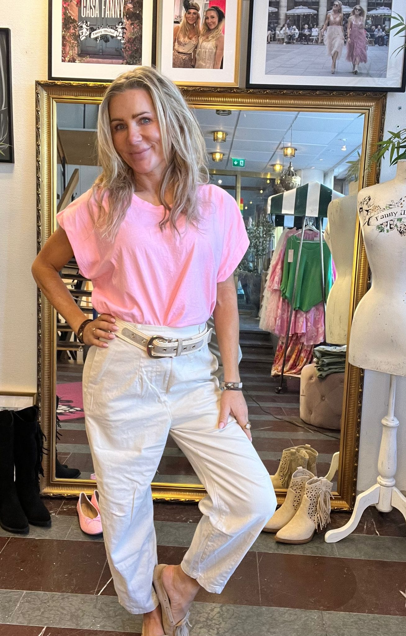 Kvinna i rosa T-shirt och vita byxor poserar leende med handen på höften framför en stor guldramsspegel i en klädbutik; klädställning och skyltdocka syns. Inramade foton på väggen, text: “CASA FANNY”.
