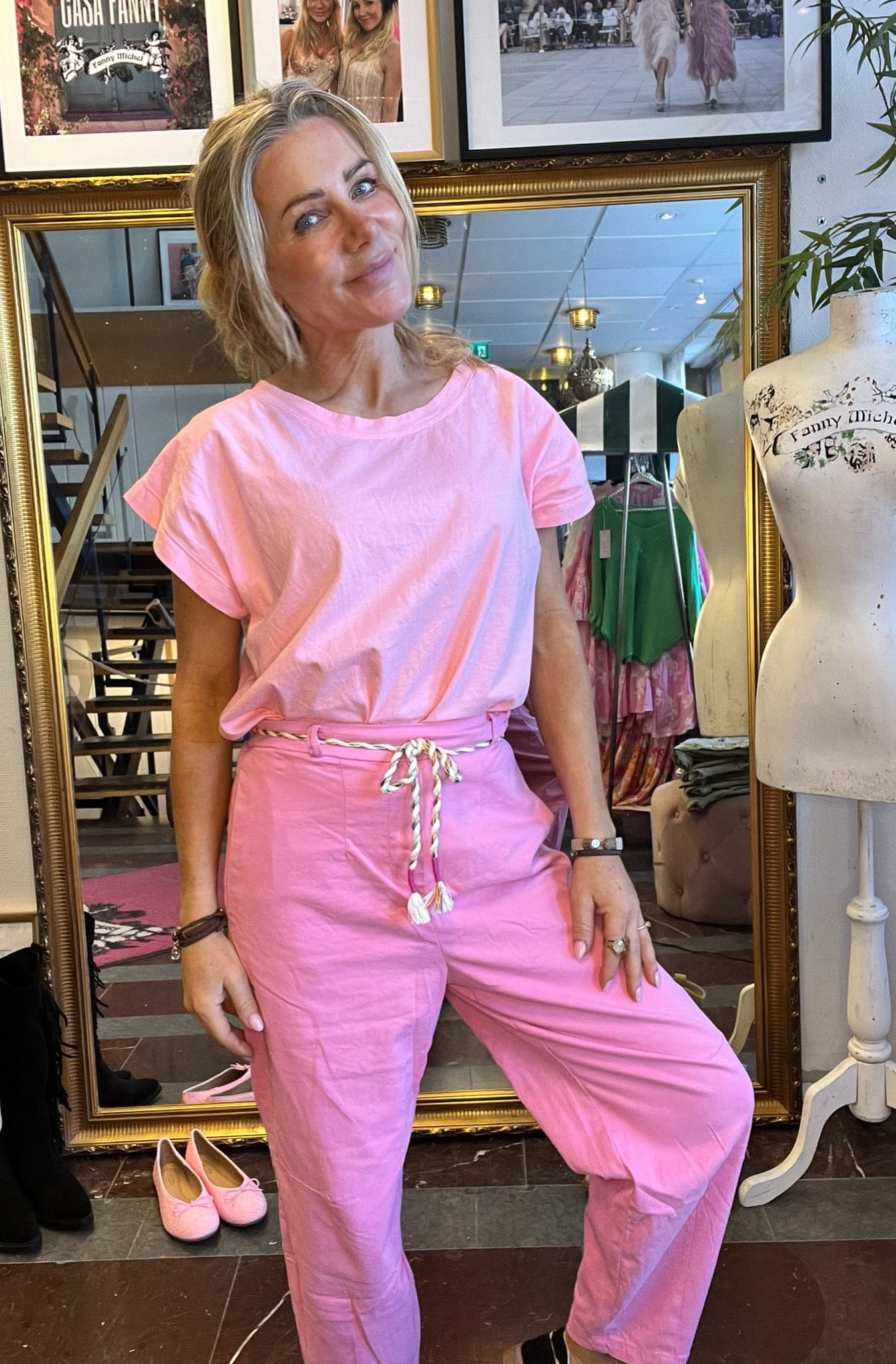 Leende kvinna poserar framför en stor guldramsspegel i en klädbutik, klädd i rosa T-shirt och rosa byxor med tvinnat snöre. Intill står en provdocka med texten ”Fanny Michel”, och rosa skor ligger på golvet.